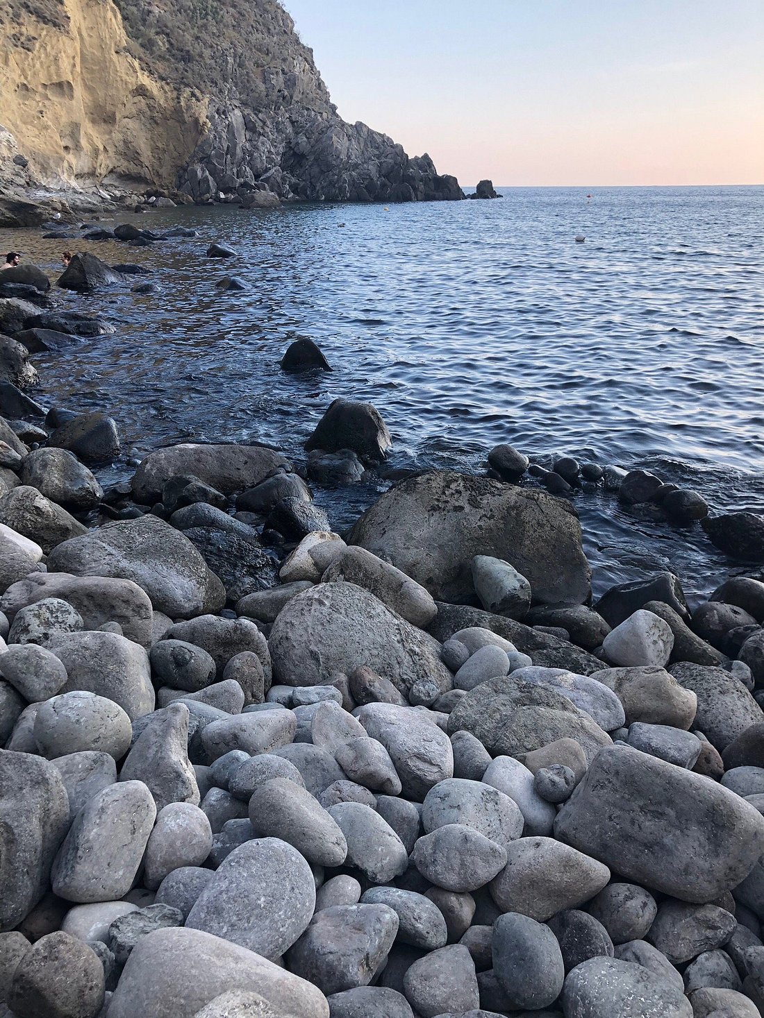 Rocky shoreline at Baia di Sorgeto in Ischia, Italy, with calm sea waters and steep cliffs in the background.