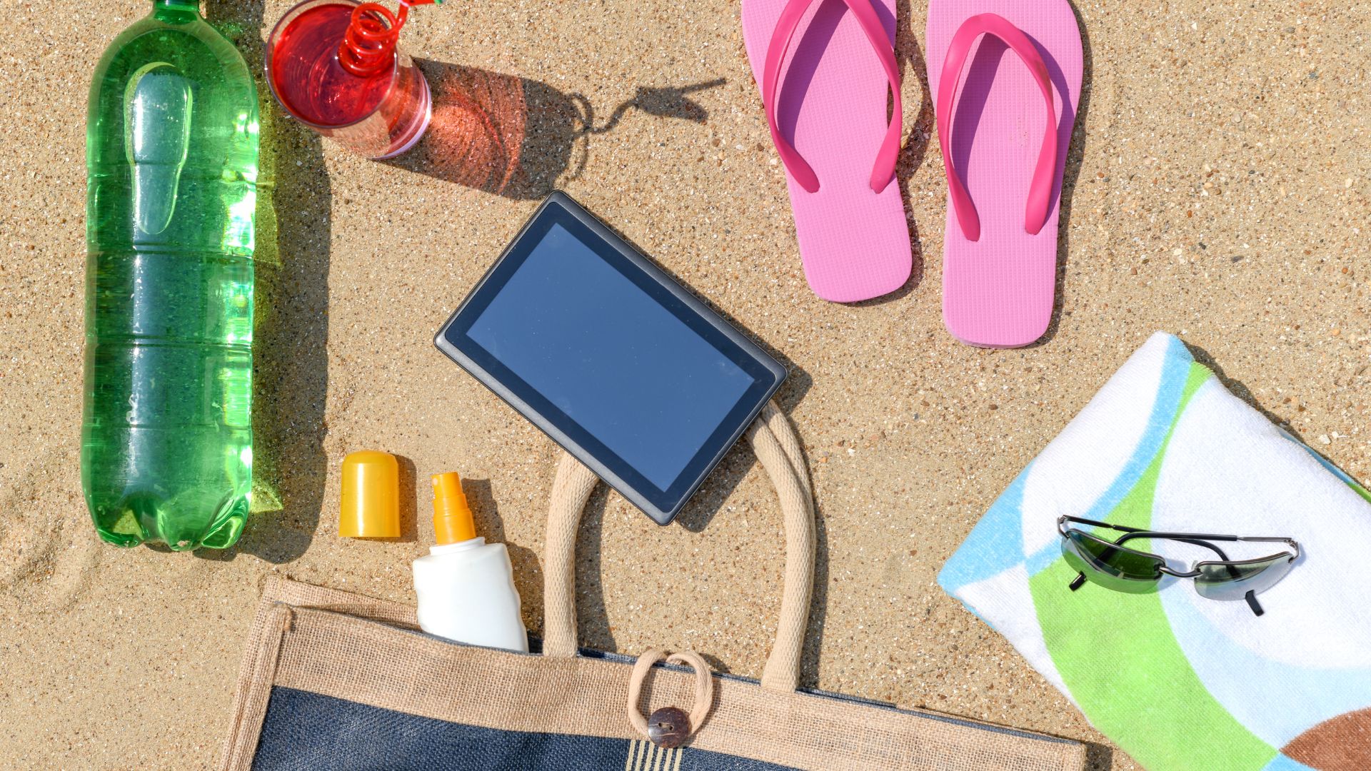 Beach bag essentials on sand, including a tablet, sunscreen, drink, and sunglasses.