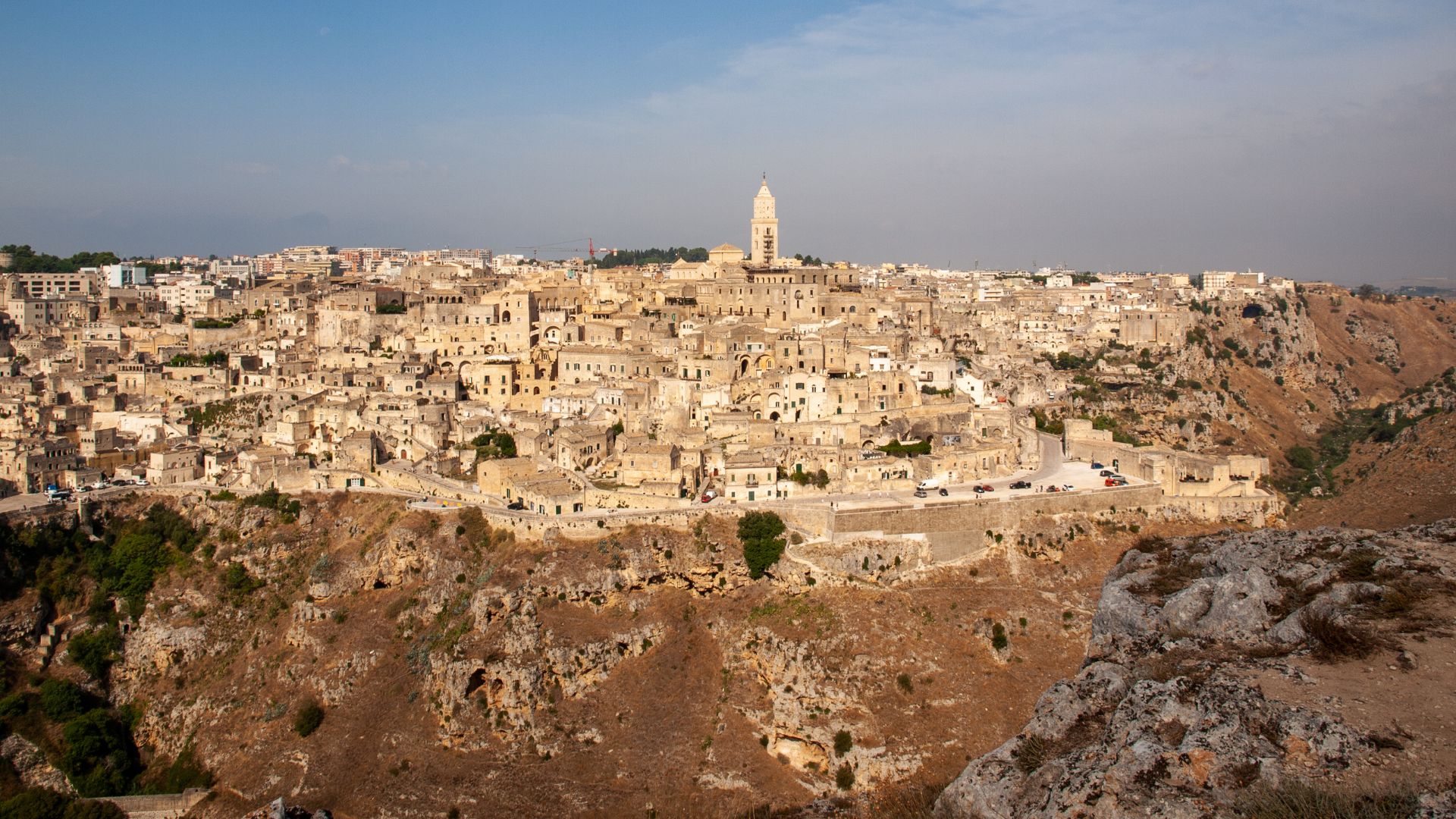 Image shows Belvedere di Murgia Timone, Sassi di Matera, Basilicata, Italy characterized by cave dwellings carved into the rock, stacked upon each other, and surrounded by a rocky landscape with sparse vegetation.