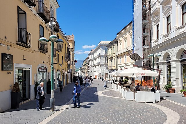 Pedestrian street in Benevento, Italy, lined with historic buildings, outdoor cafes, and people enjoying a sunny day.