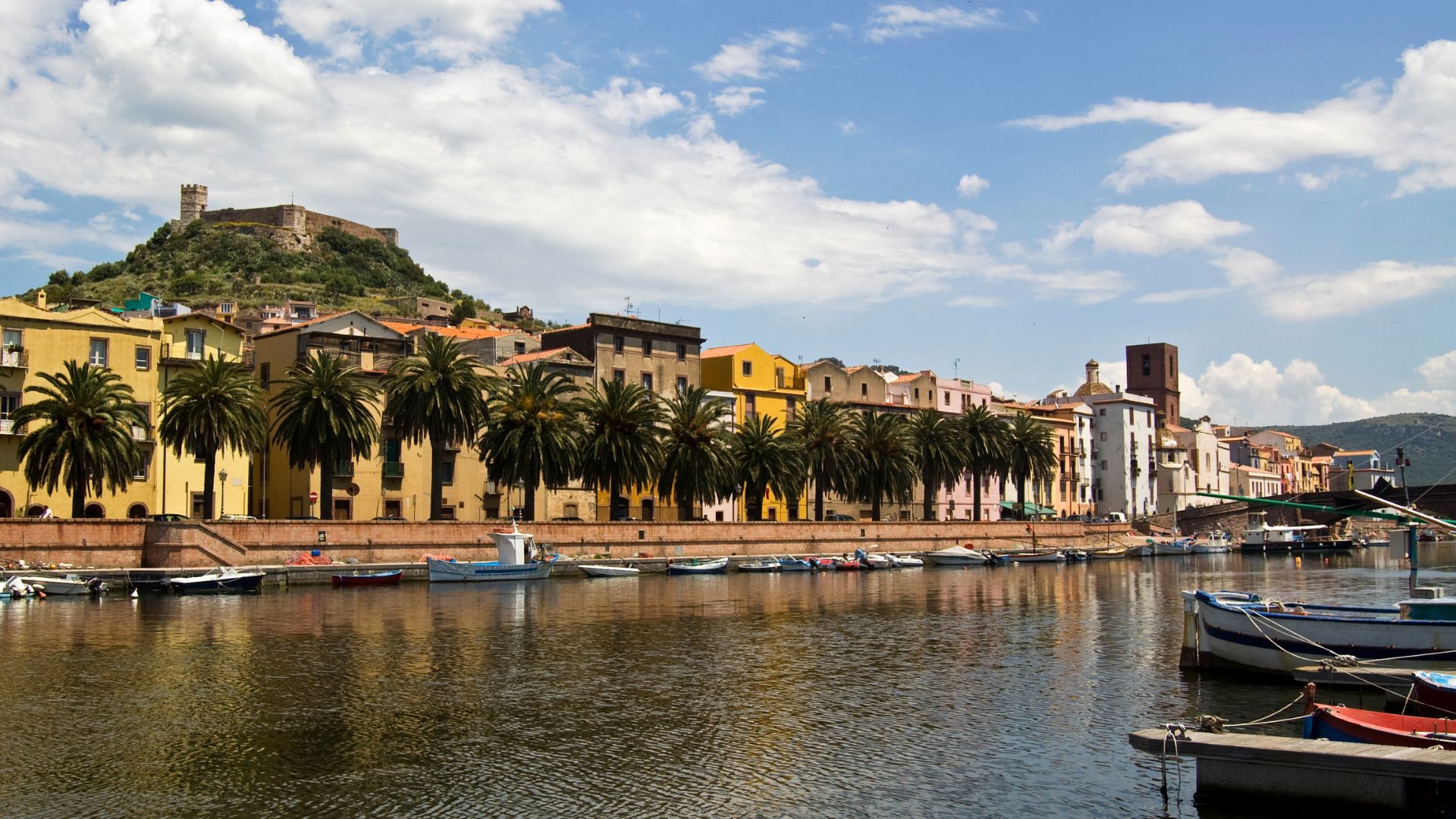 Bosa, Italy, is depicted with colorful buildings lining the Temo River, boats on the water, and a castle on the hilltop in the background. The scene is set under a partly cloudy sky.