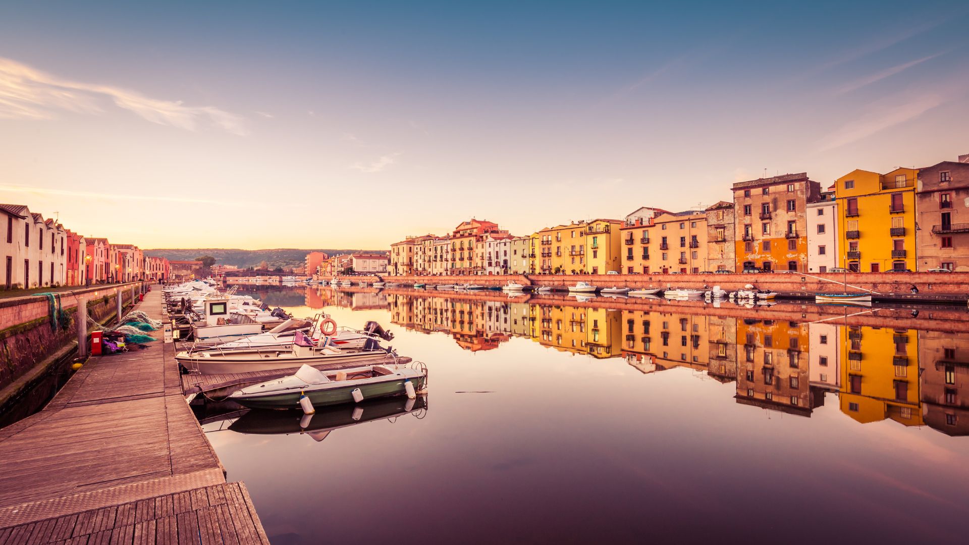 Bosa's town in Sardinia, Italy, is characterized by its colorful buildings lining the Temo River, and some bots on dock with reflections visible in the water, under a clear blue sky.