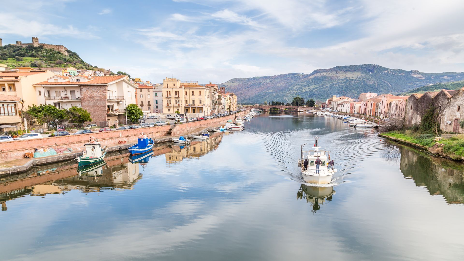 Bosa, Italy, is depicted with colorful buildings lining the Temo River, boats on the water, and a castle on the hilltop in the background.