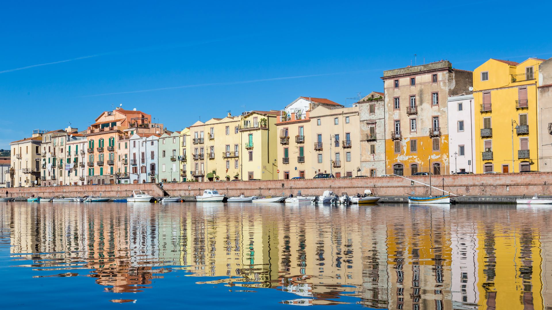 Bosa's town in Sardinia, Italy, is characterized by its colorful buildings lining the Temo River, with reflections visible in the water, under a clear blue sky.