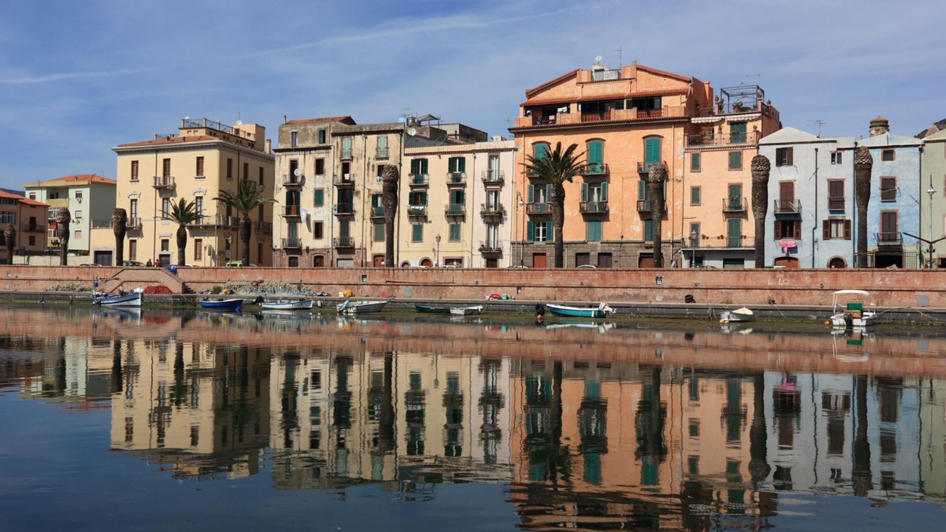 Bosa's town in Sardinia, Italy, is characterized by its colorful buildings lining the Temo River, with reflections visible in the water, under a clear blue sky.