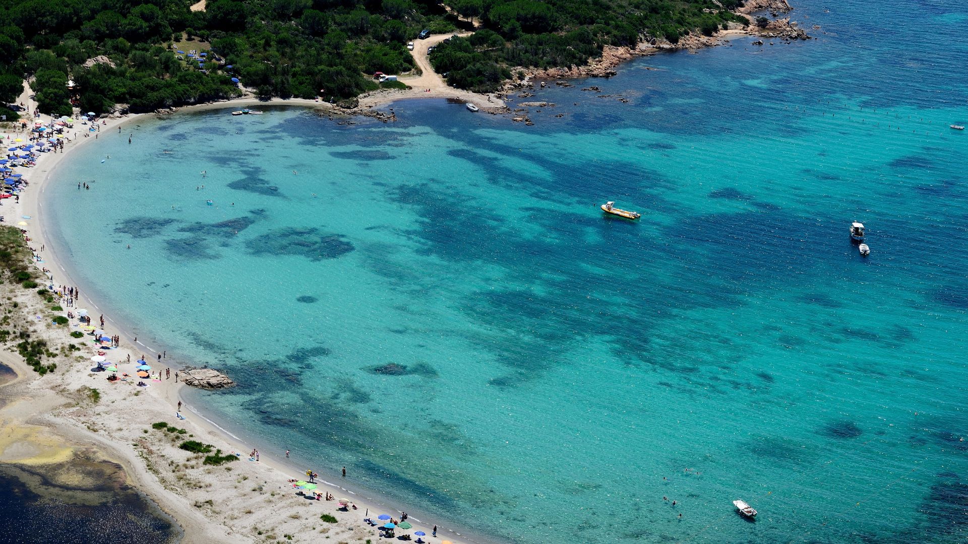 Aerial view of Cala Brandinchi, Sardinia, showing a white sandy beach, clear turquoise water, boats, and surrounding greenery.