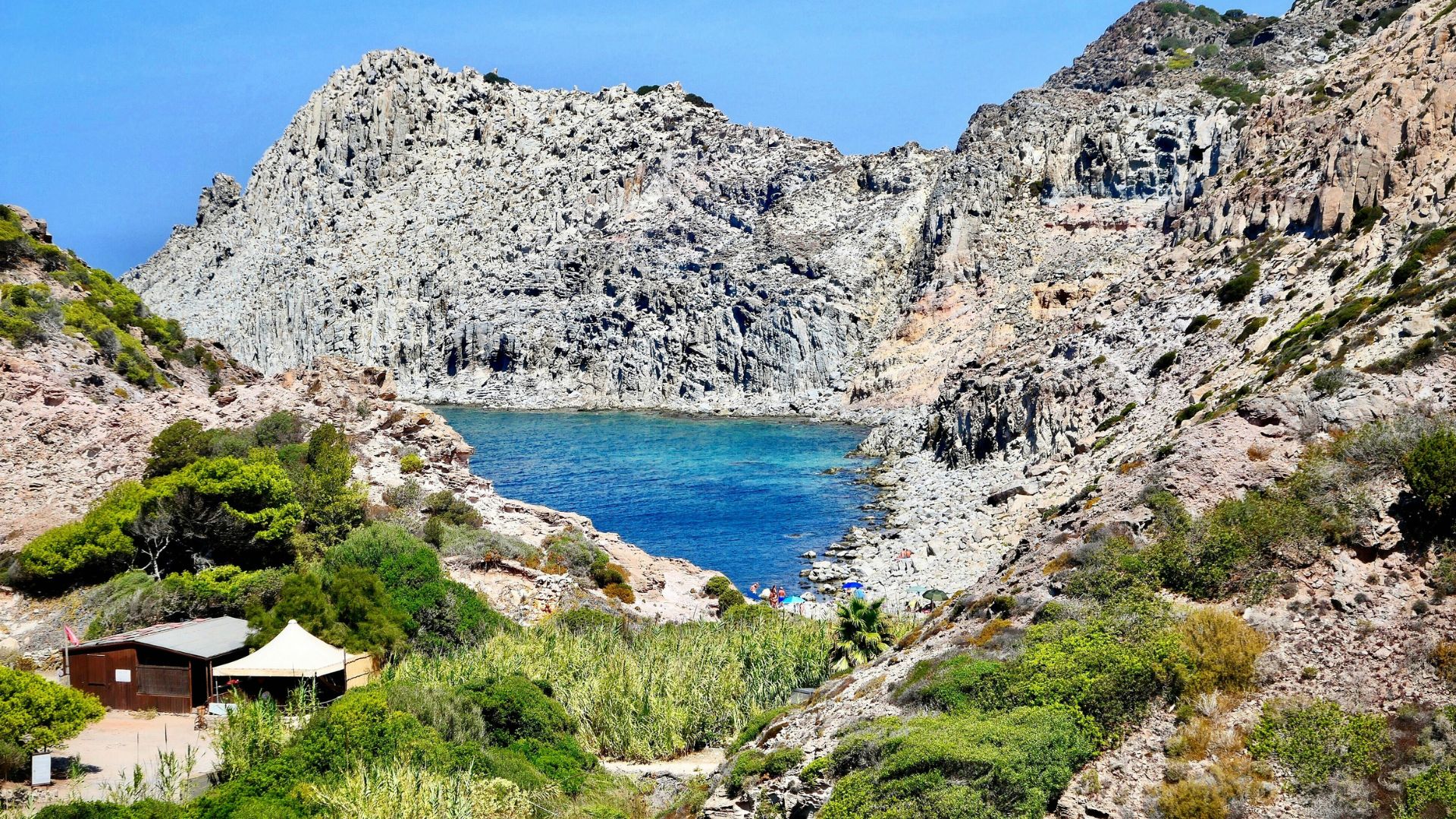 A landscape view of Cala Fico beach in Carloforte, Sardinia, showcasing a cove of turquoise water surrounded by rugged, rocky cliffs and sparse vegetation, with a small building and tent visible on the left side.