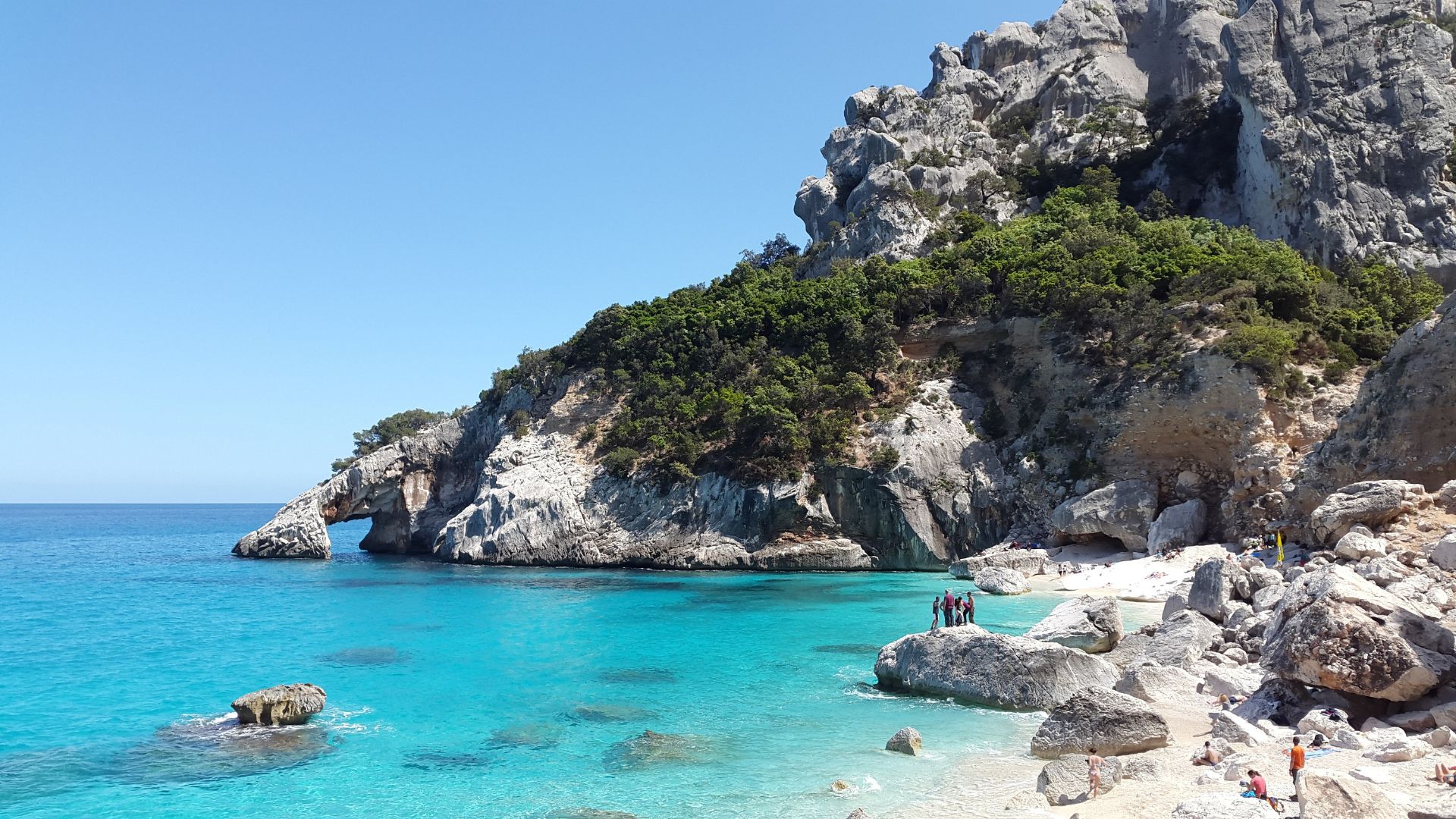 Cala Goloritzé, a picturesque beach in Sardinia, featuring turquoise waters, a white pebble beach, towering limestone cliffs, and a natural rock arch.