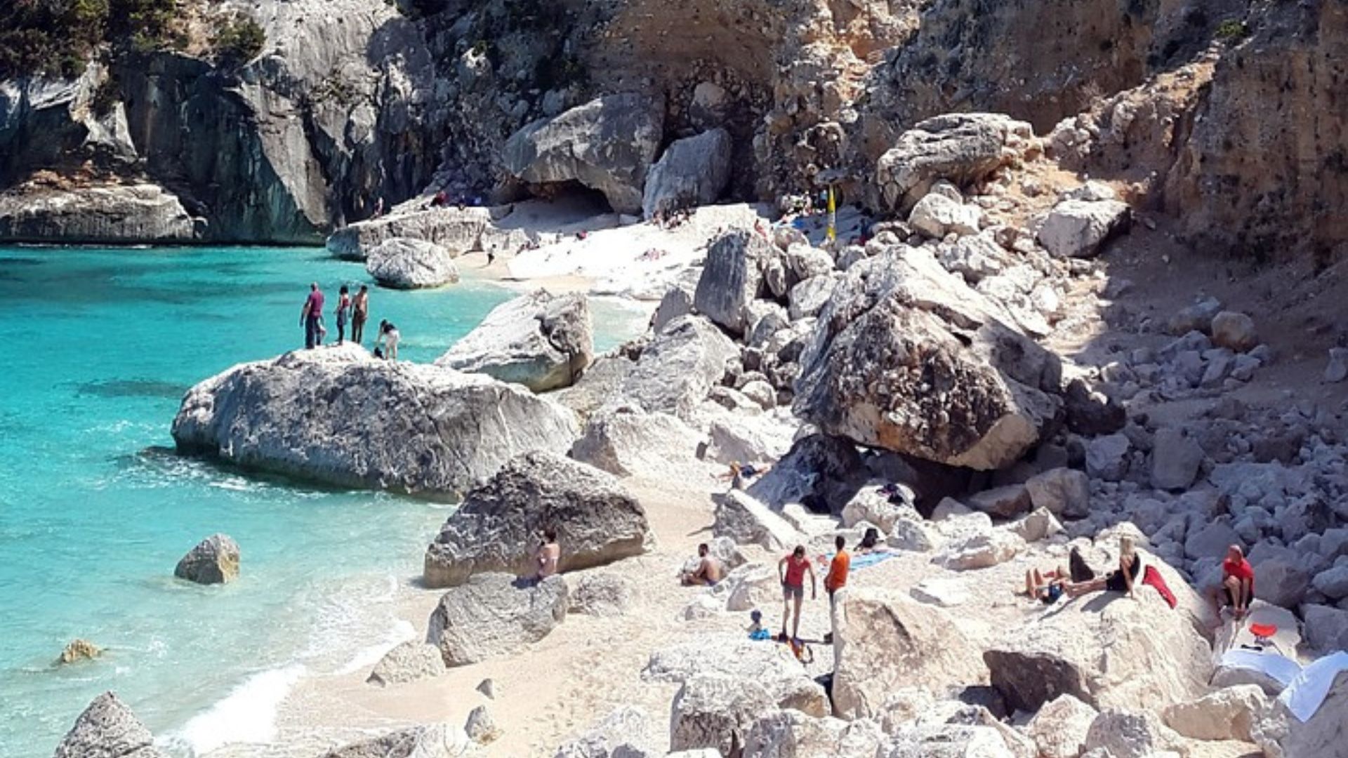  A scenic view of Cala Goloritzé beach in Sardinia, Italy, featuring turquoise water, white pebble beach, large rock formations, and people enjoying the beach