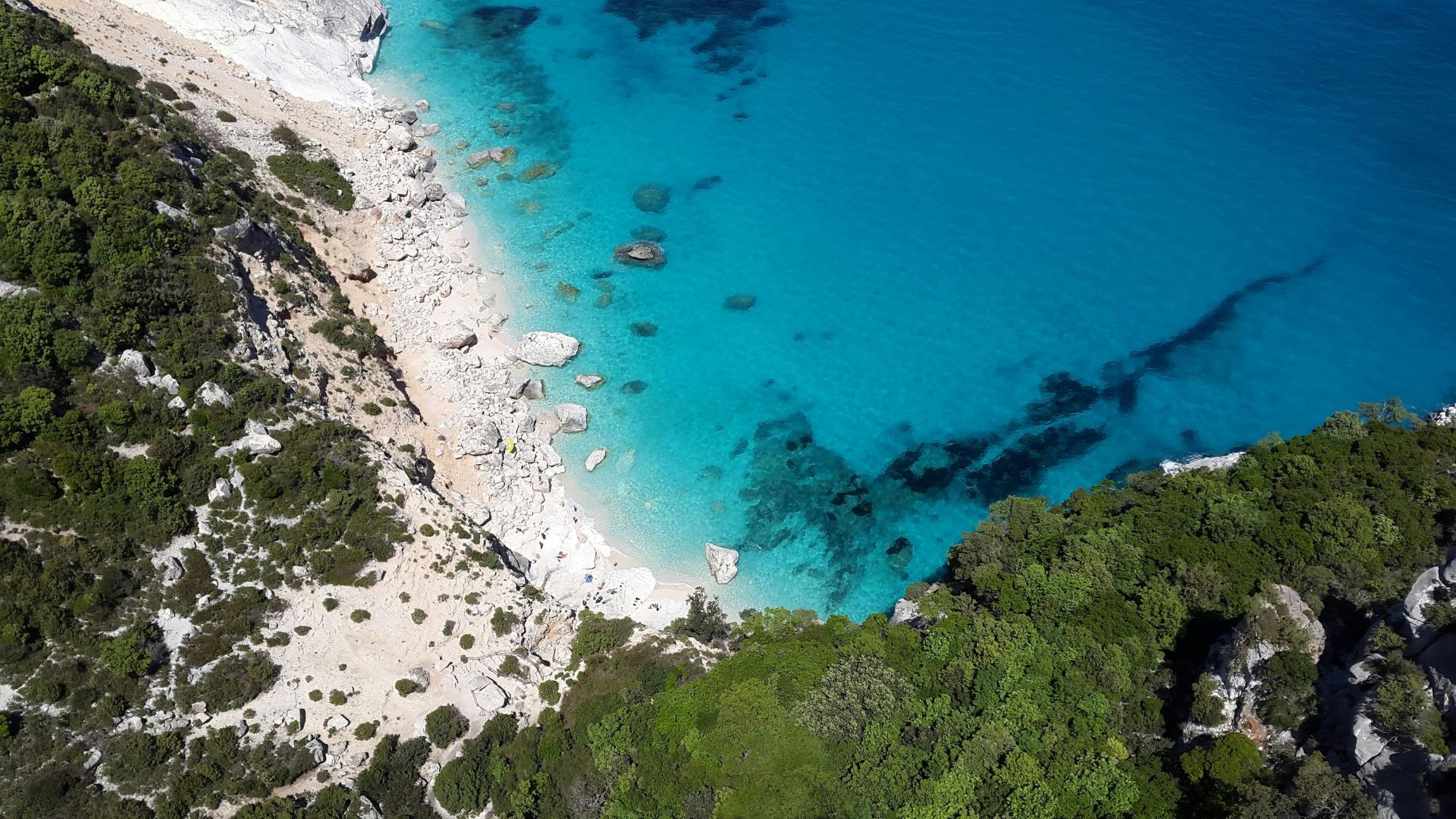 Aerial view of Cala Goloritzé, a small beach with white pebbles and turquoise water, surrounded by rocky cliffs and lush vegetation, with people relaxing on the shore.