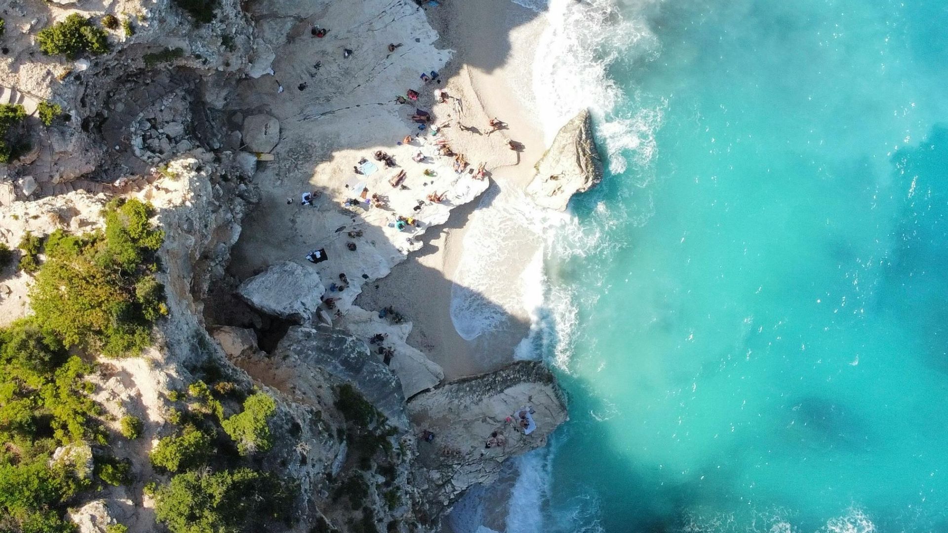 Aerial view of Cala Goloritzé, a small beach with white pebbles and turquoise water, surrounded by rocky cliffs and lush vegetation, with people relaxing on the shore.