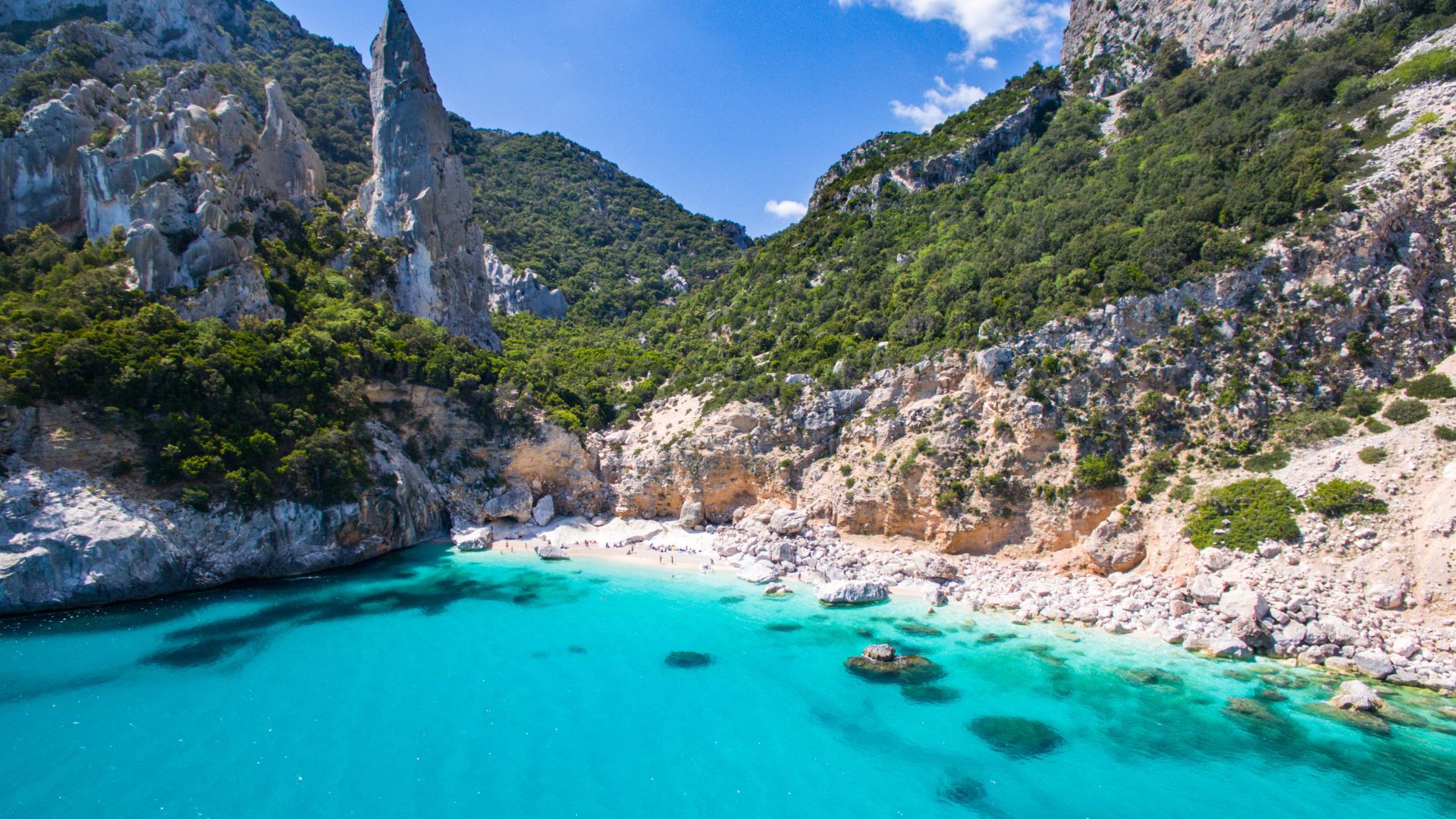 Aerial view of Cala Goloritzé in Sardinia, featuring turquoise waters, a white pebble beach, towering limestone cliffs, and a prominent pinnacle.