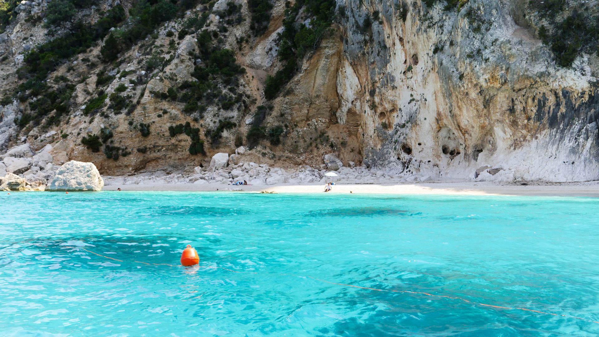 A scenic view of Cala Gonone in Sardinia, featuring turquoise water, a sandy beach, and rocky cliffs covered with vegetation. A red buoy is visible in the foreground, and the coastline stretches into the distance.