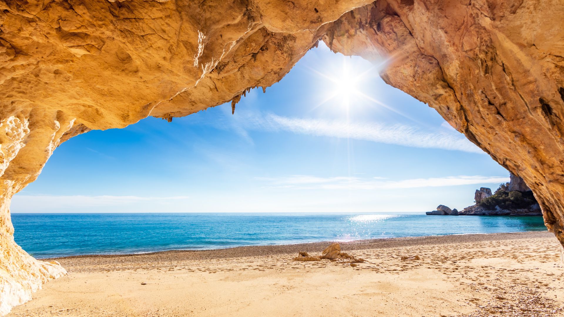 View from a cave entrance overlooking Cala Luna beach at sunset, with boats on the water and a colorful sky.