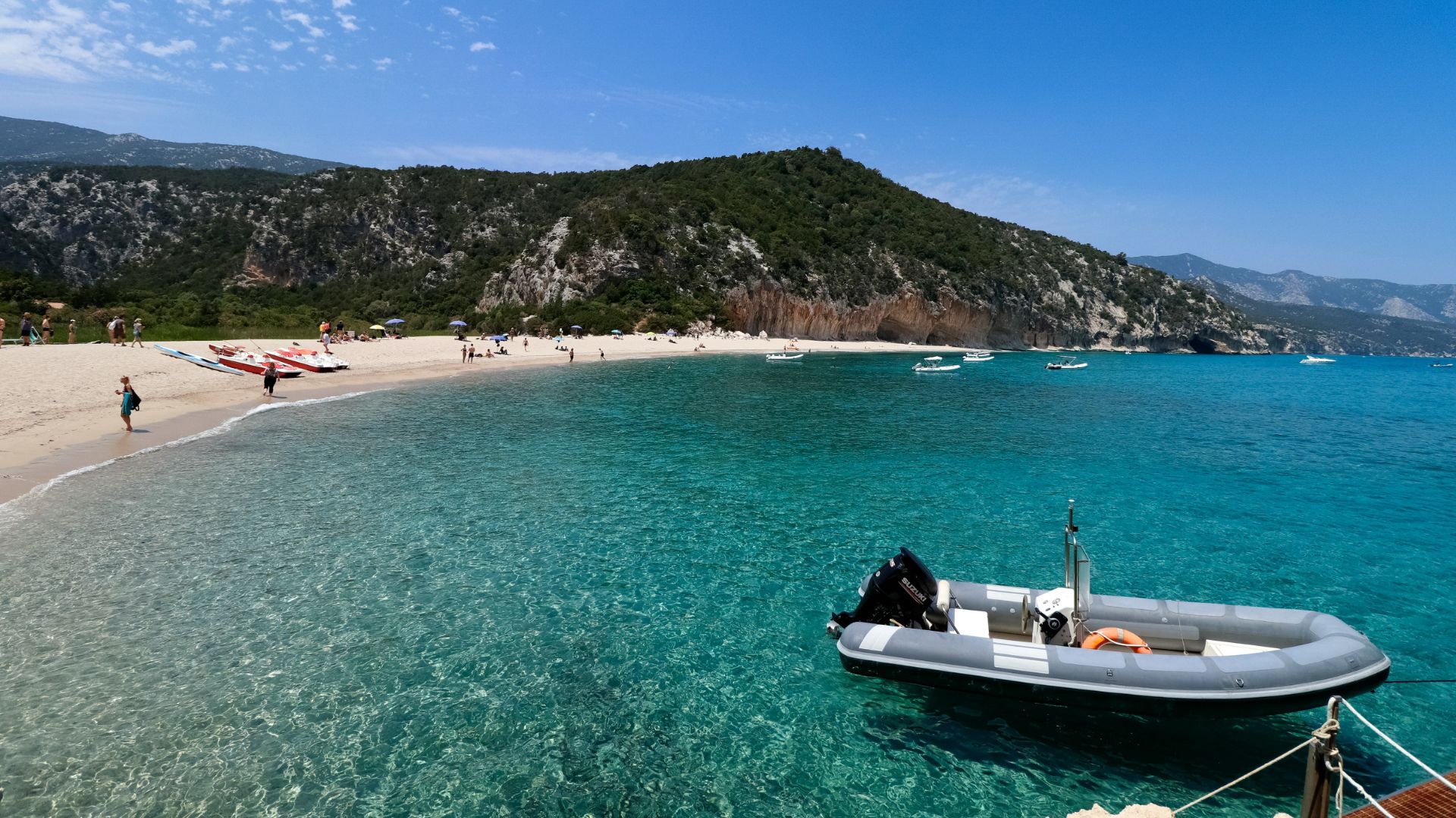 A scenic view of Cala Luna beach in Sardinia, featuring a crescent-shaped sandy beach, clear turquoise water, a small boat floating in the foreground, and a backdrop of rocky cliffs and green vegetation under a clear blue sky.
