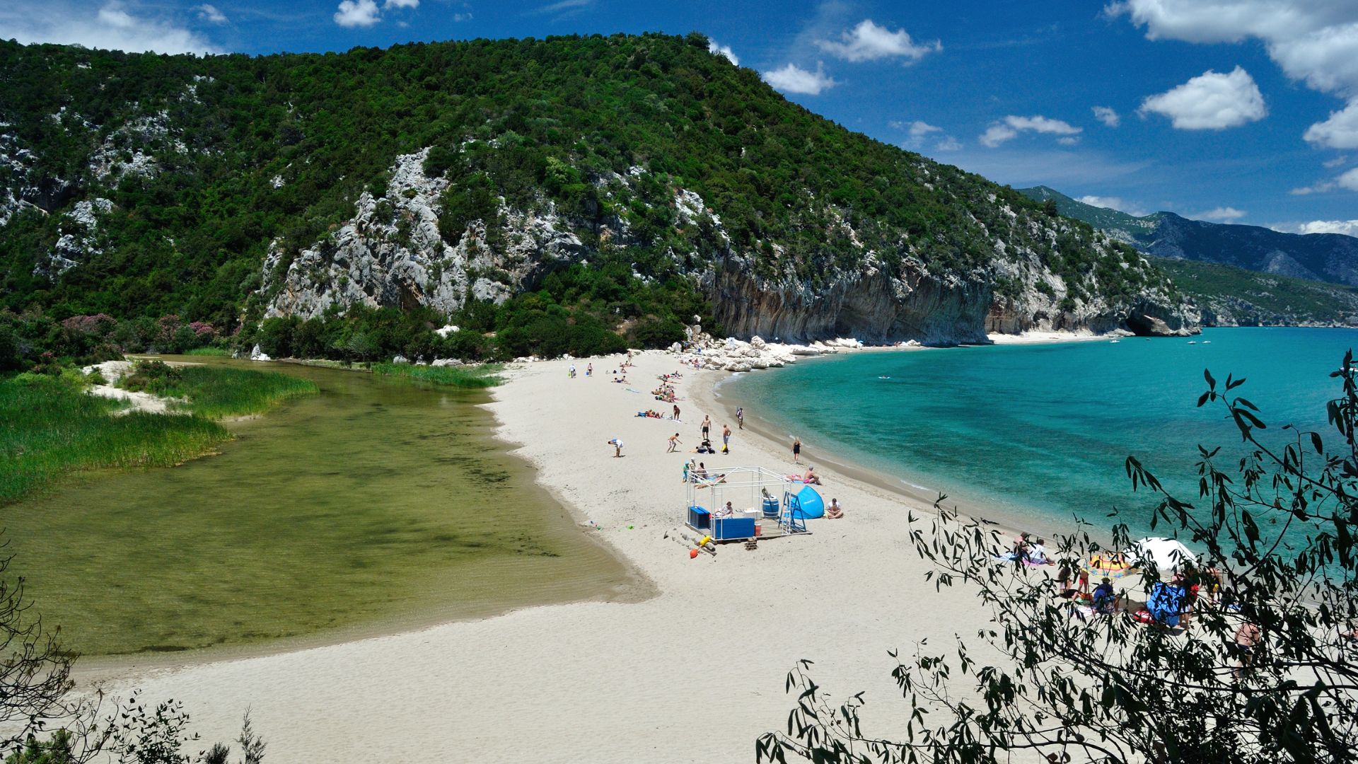 A picturesque beach scene with clear turquoise water, white sand, and towering, rugged cliffs. The cove is framed by natural caves and lush greenery, with the sun shining brightly above. 