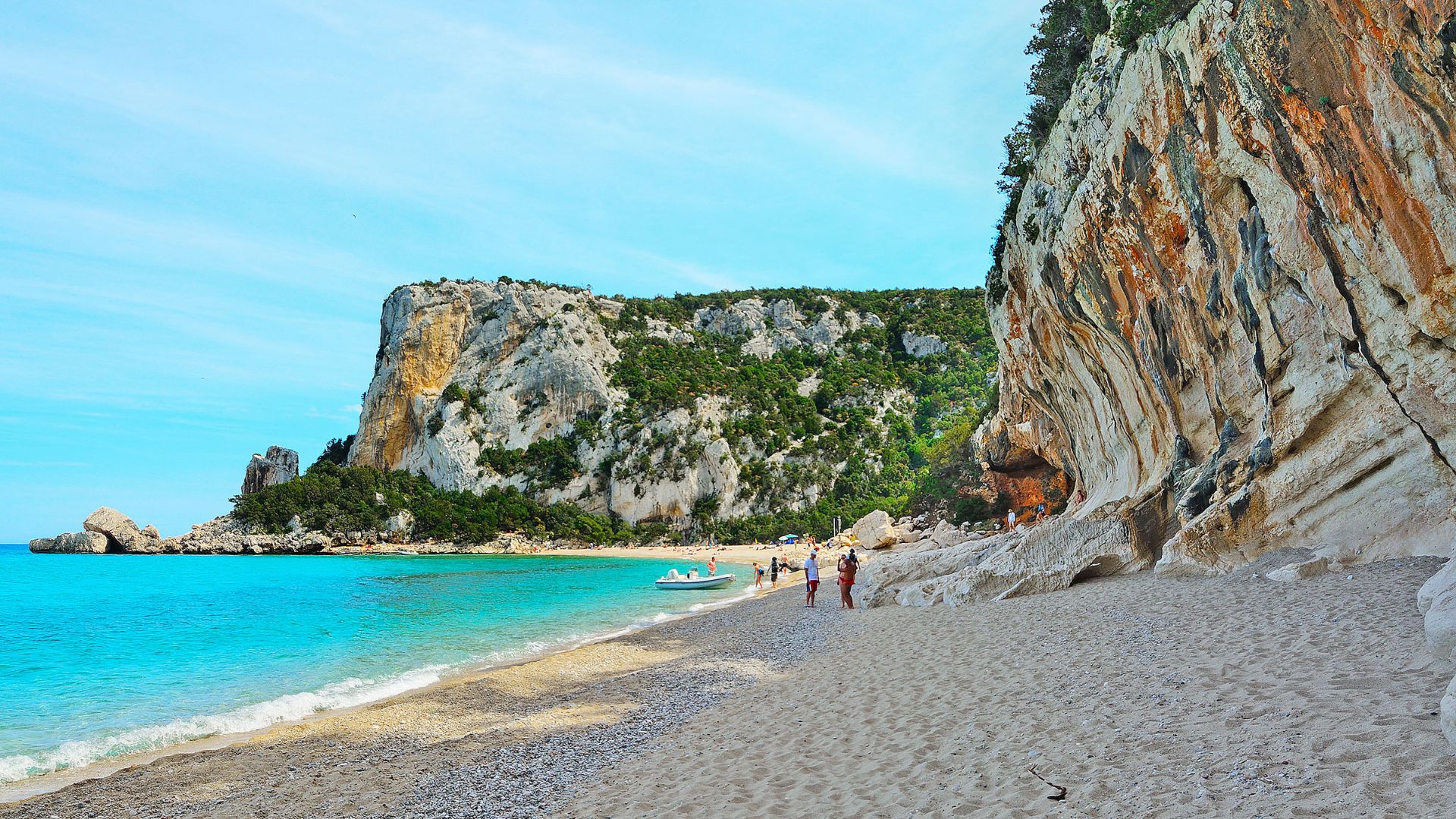 Panoramic view of Cala Luna beach in Sardinia, Italy, showing the crescent-shaped beach, turquoise waters, limestone cliffs with caves, and surrounding green hills under a clear blue sky.