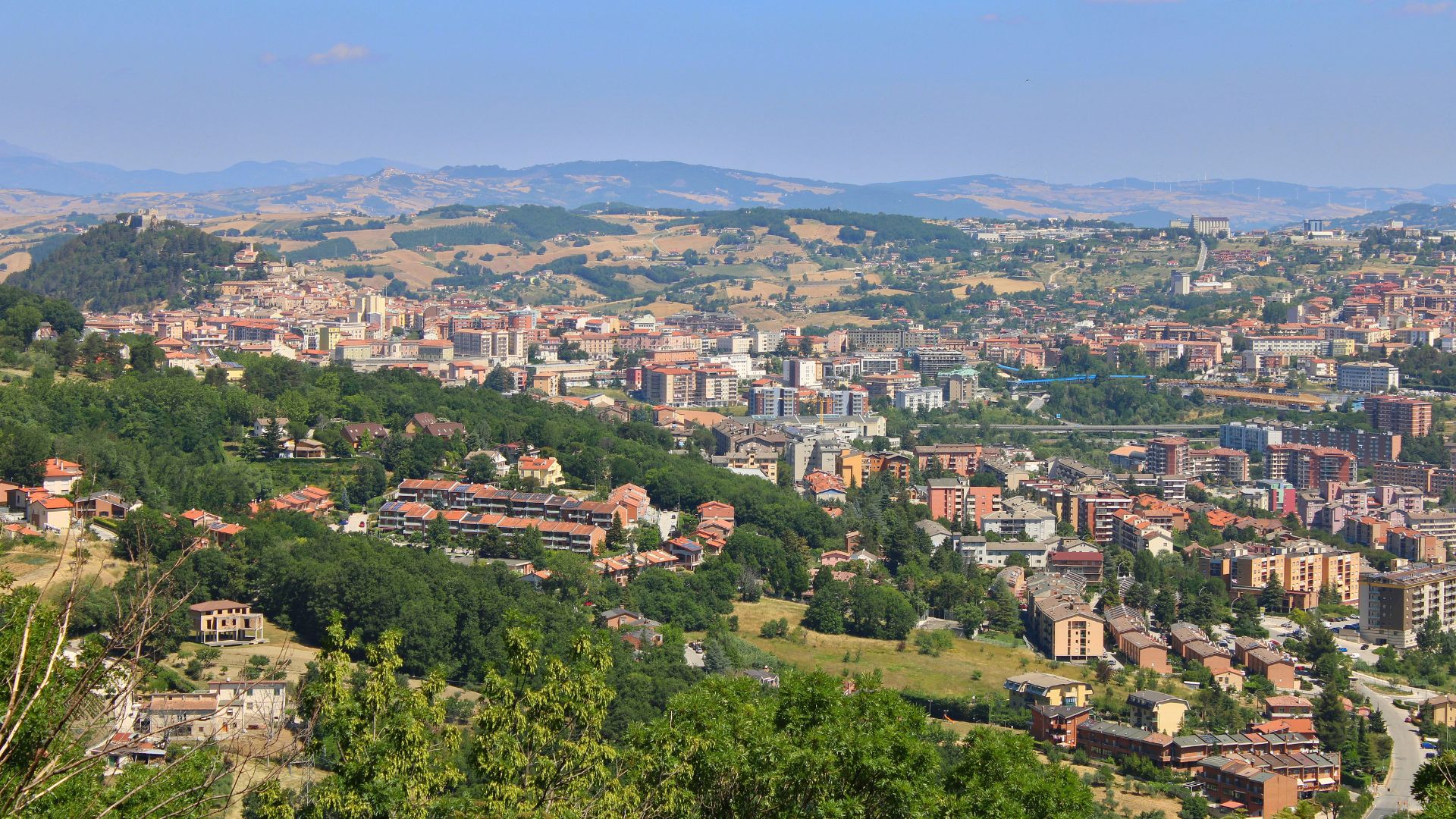 A hilltop view captures a densely populated town with buildings of varying heights and colors, nestled amidst lush green trees and rolling hills under a clear blue sky. Distant mountains are visible on the horizon.