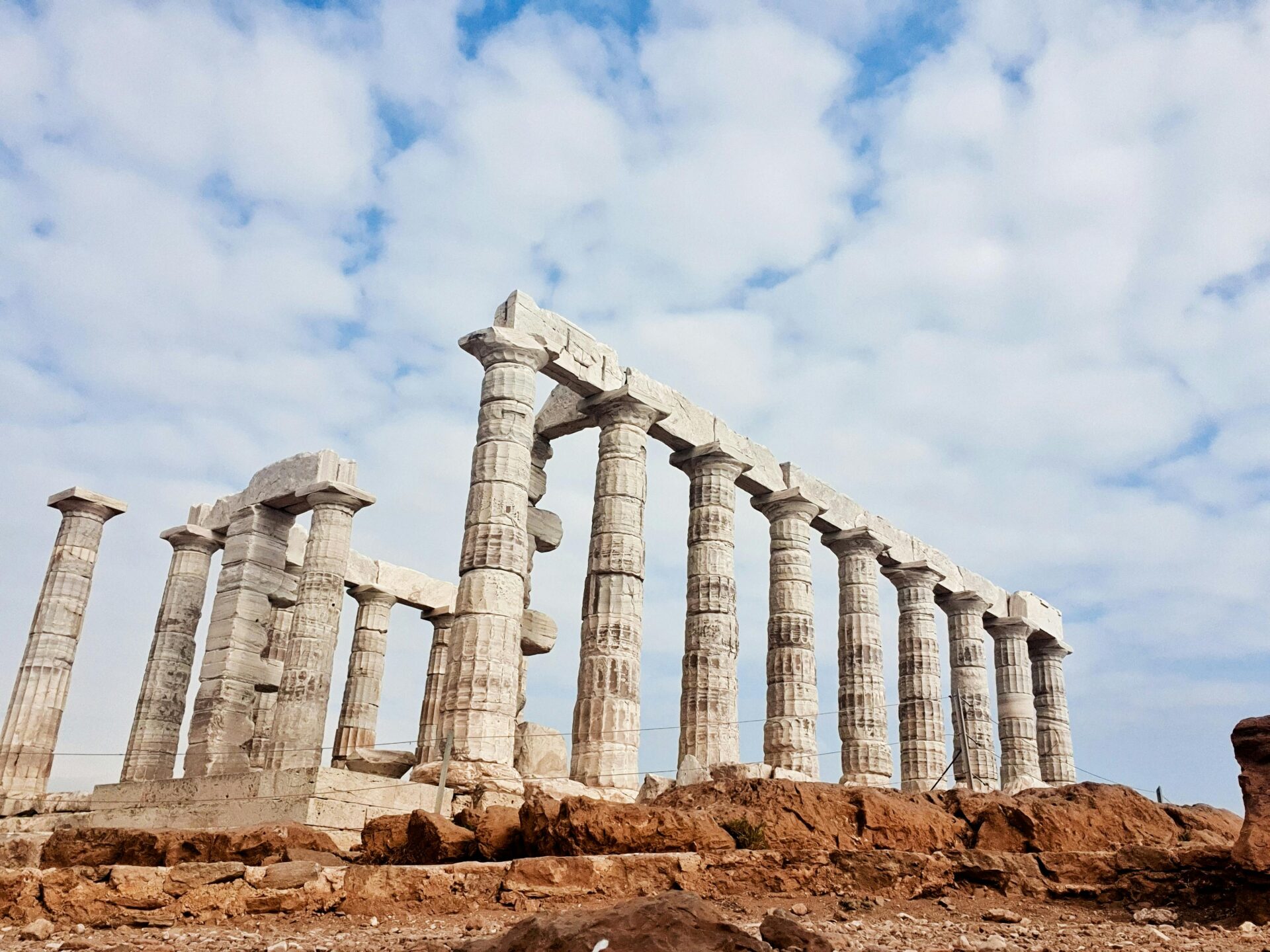 Ruins of the ancient Temple of Poseidon at Cape Sounion, Greece, standing on a rocky cliff under a partly cloudy sky.