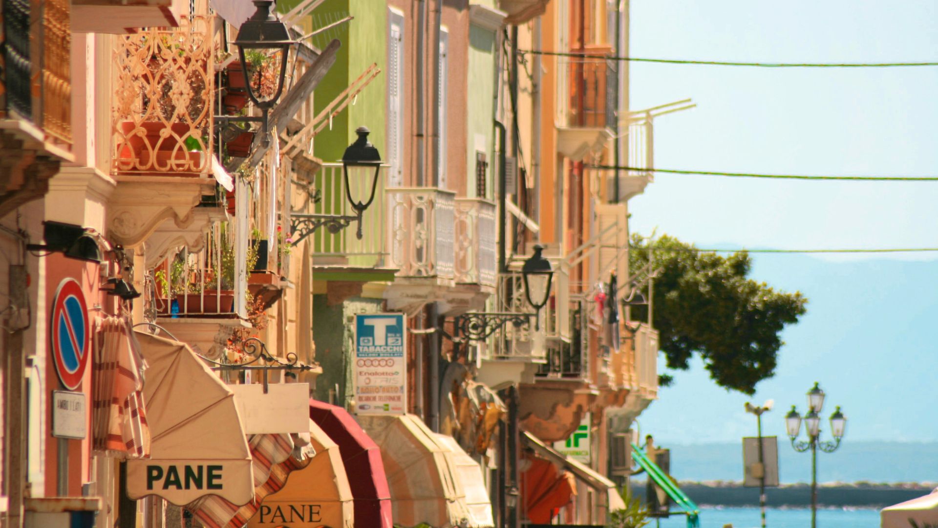 Colorful buildings line a street in Carloforte, Sardinia, Italy, with awnings shading shop fronts and the sea visible in the background. Balconies with ornate railings and decorative street lamps add to the charm of the scene. Signs with "PANE" are visible on the awnings, indicating a bakery or bread shop.