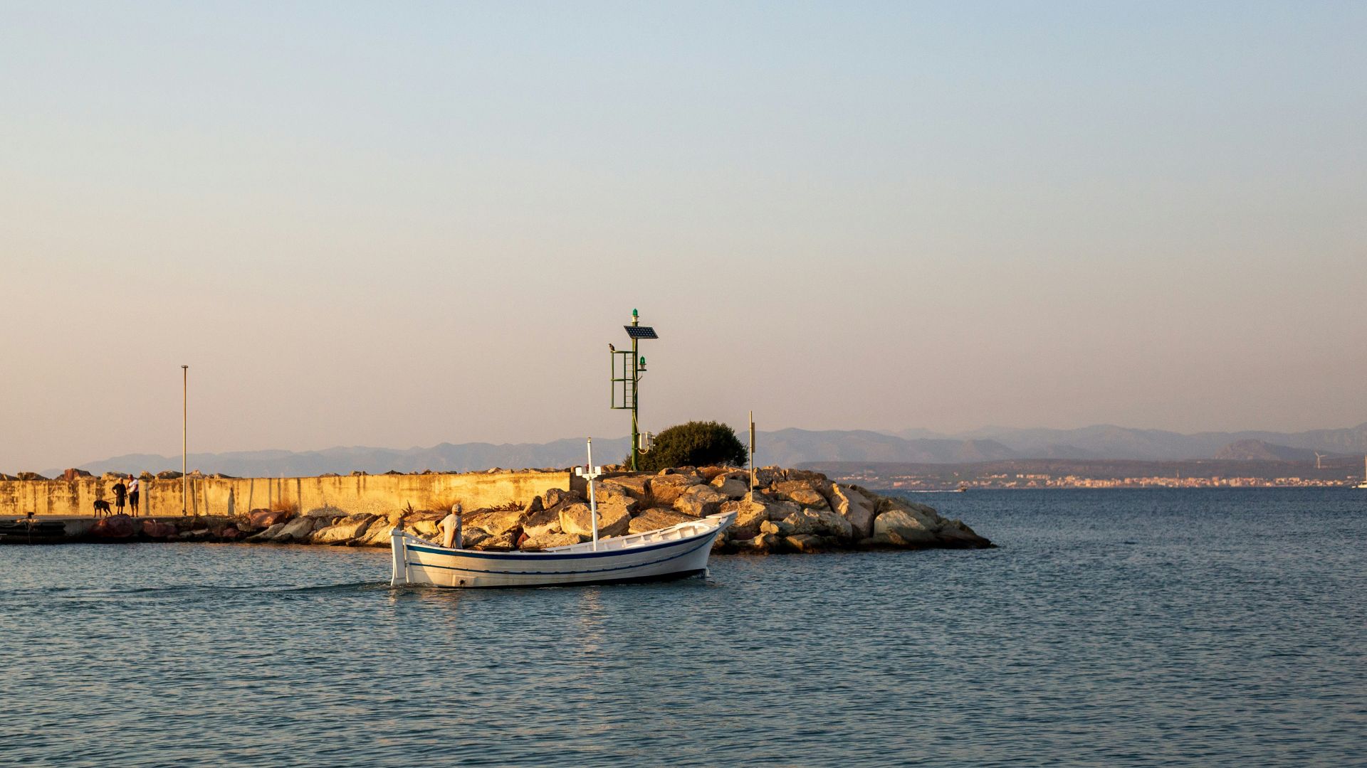 A white fishing boat navigates calm waters near a rocky breakwater and navigation beacon in Carloforte, Sardinia.