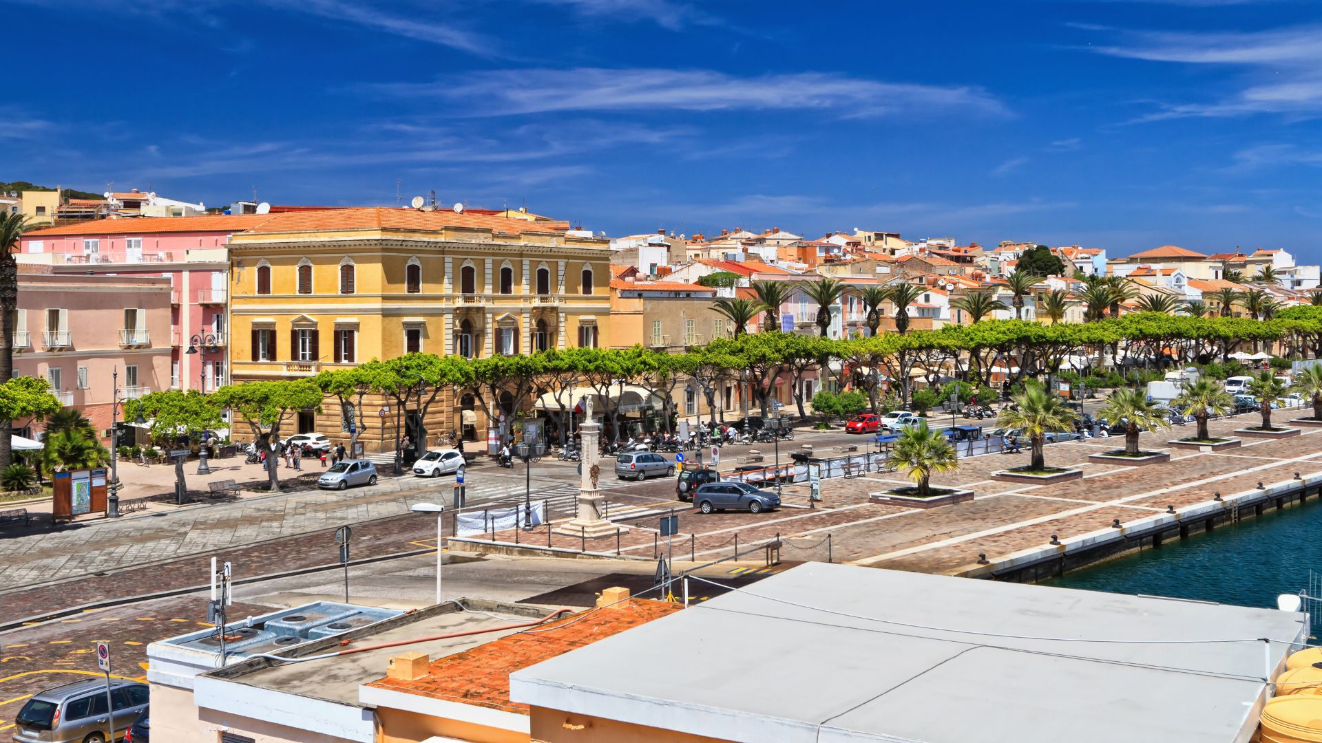A high-angle, wide shot depicts a coastal town with colorful buildings, palm trees, and parked cars, set against a clear blue sky. 