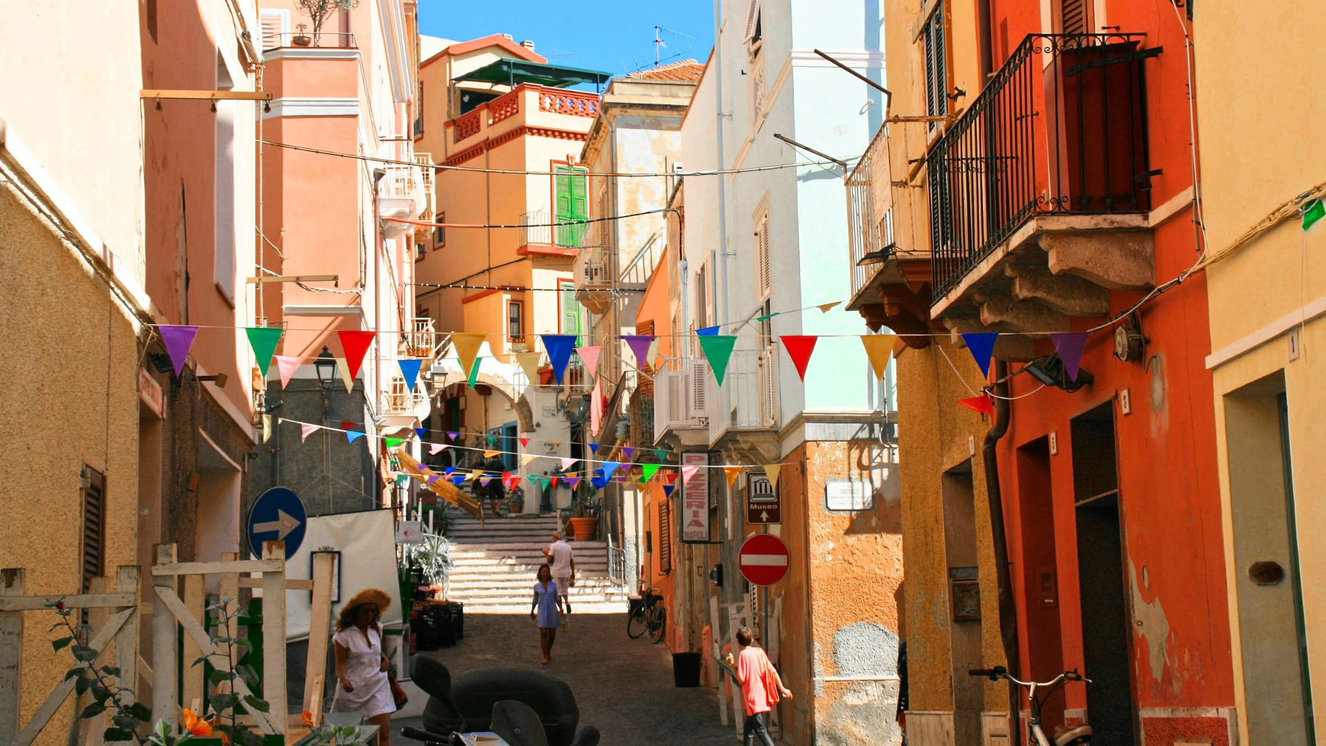 A narrow street in Carloforte, Sardinia, Italy decorated with colorful bunting flags, with buildings in shades of pink, orange, and white on either side, and people walking in the distance. A blue directional sign and a red and white no-entry sign are visible on the left and right sides of the street, respectively.