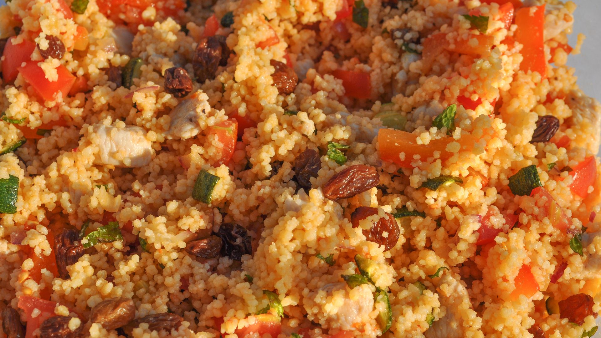  A close-up shot of couscous mixed with vegetables and raisins. The couscous is the predominant element, with visible pieces of tomato, green herbs, and scattered raisins throughout.