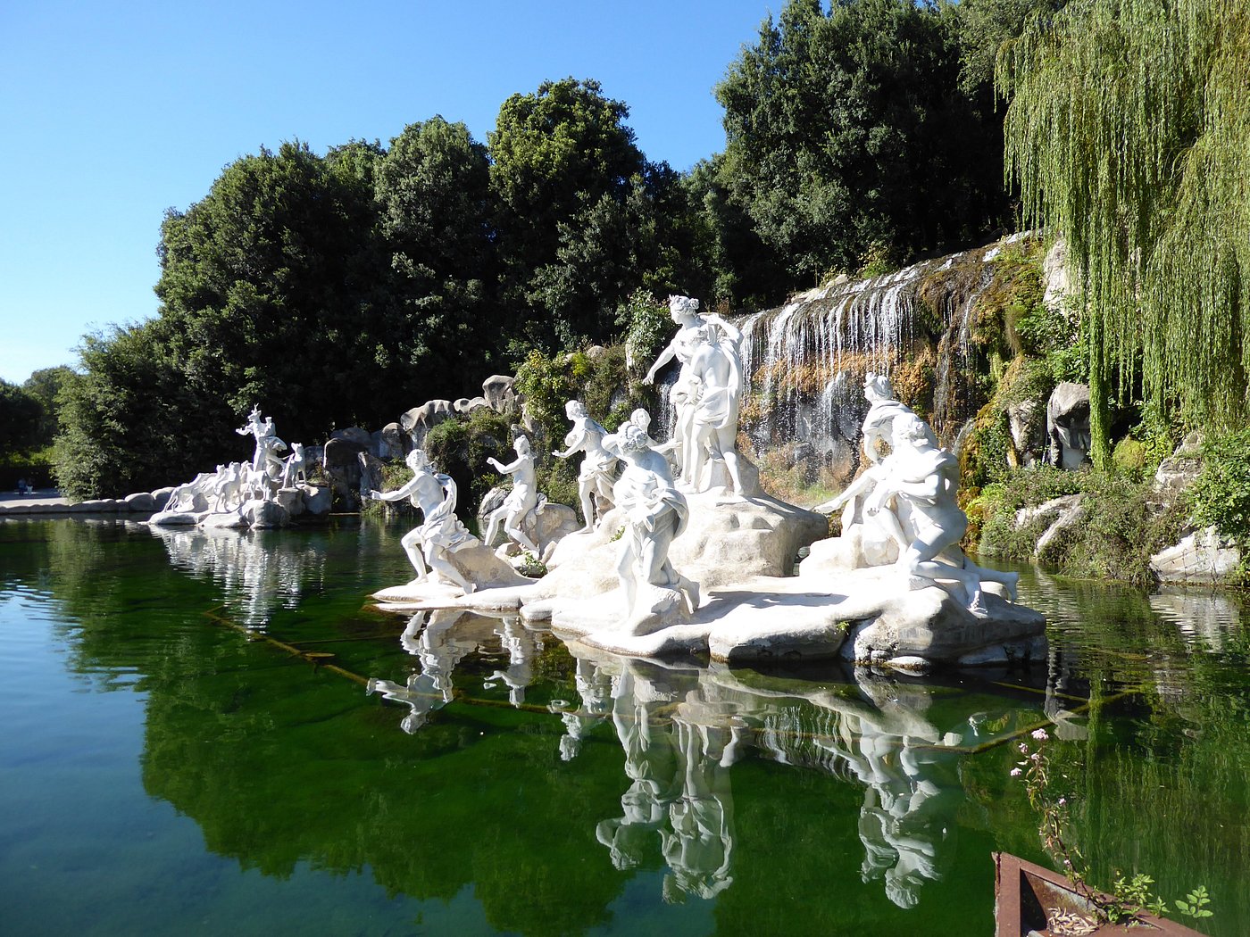 White marble statues by a waterfall and reflective pond in Caserta’s Royal Park, Italy.