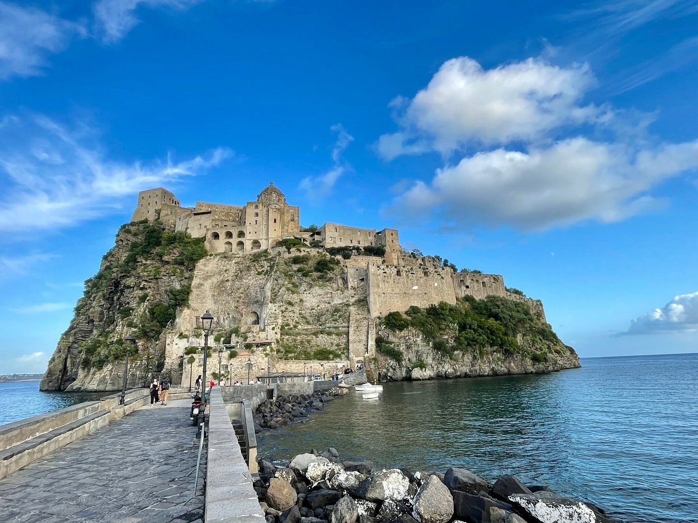View of Castello Aragonese on a rocky islet connected to Ischia by a stone bridge, under a bright blue sky.