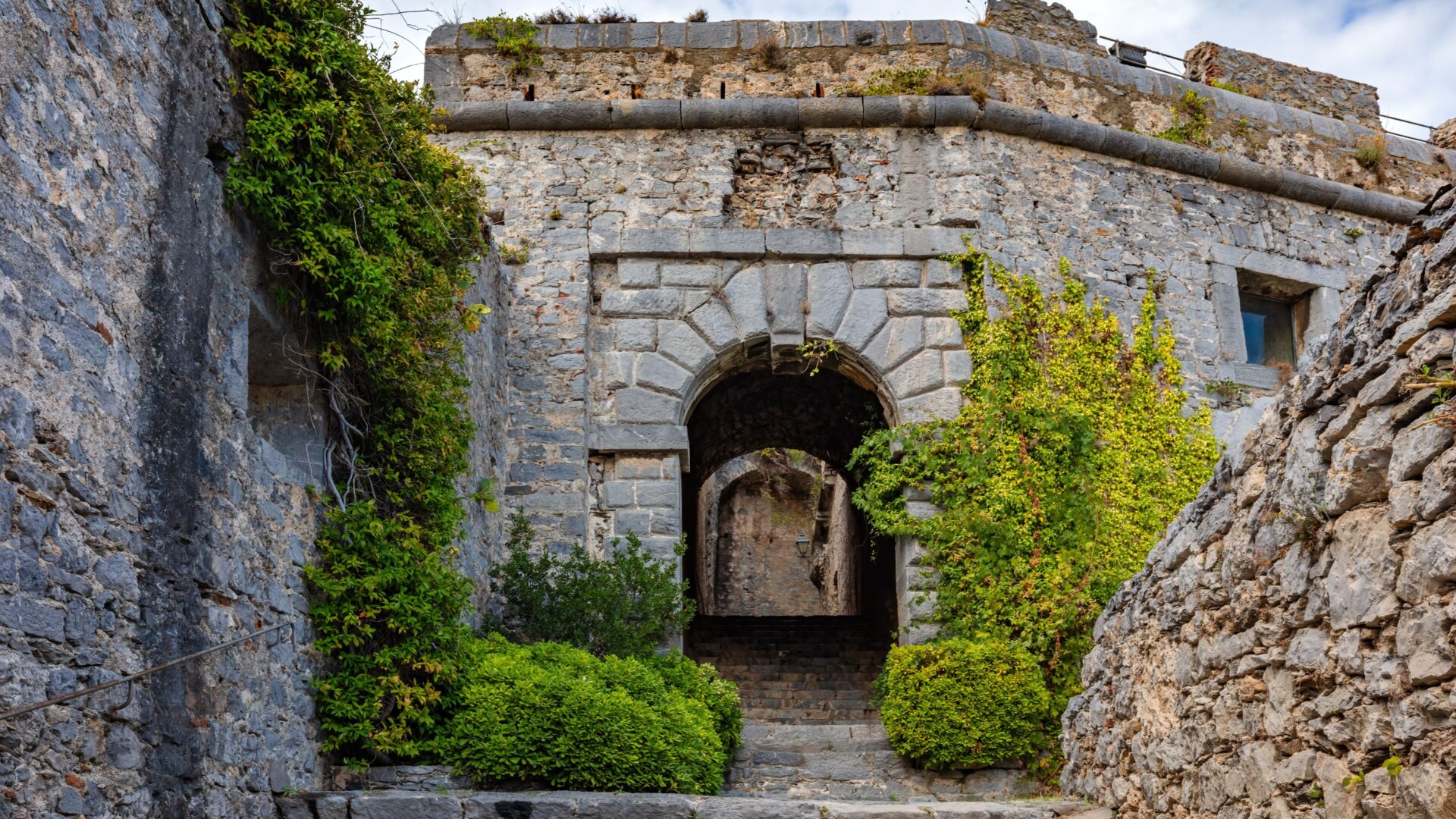 A stone castle, Castello Doria, sits atop a hill in Castelsardo, Sardinia, surrounded by green vegetation and stone walls, with an arched entrance leading into the castle.
