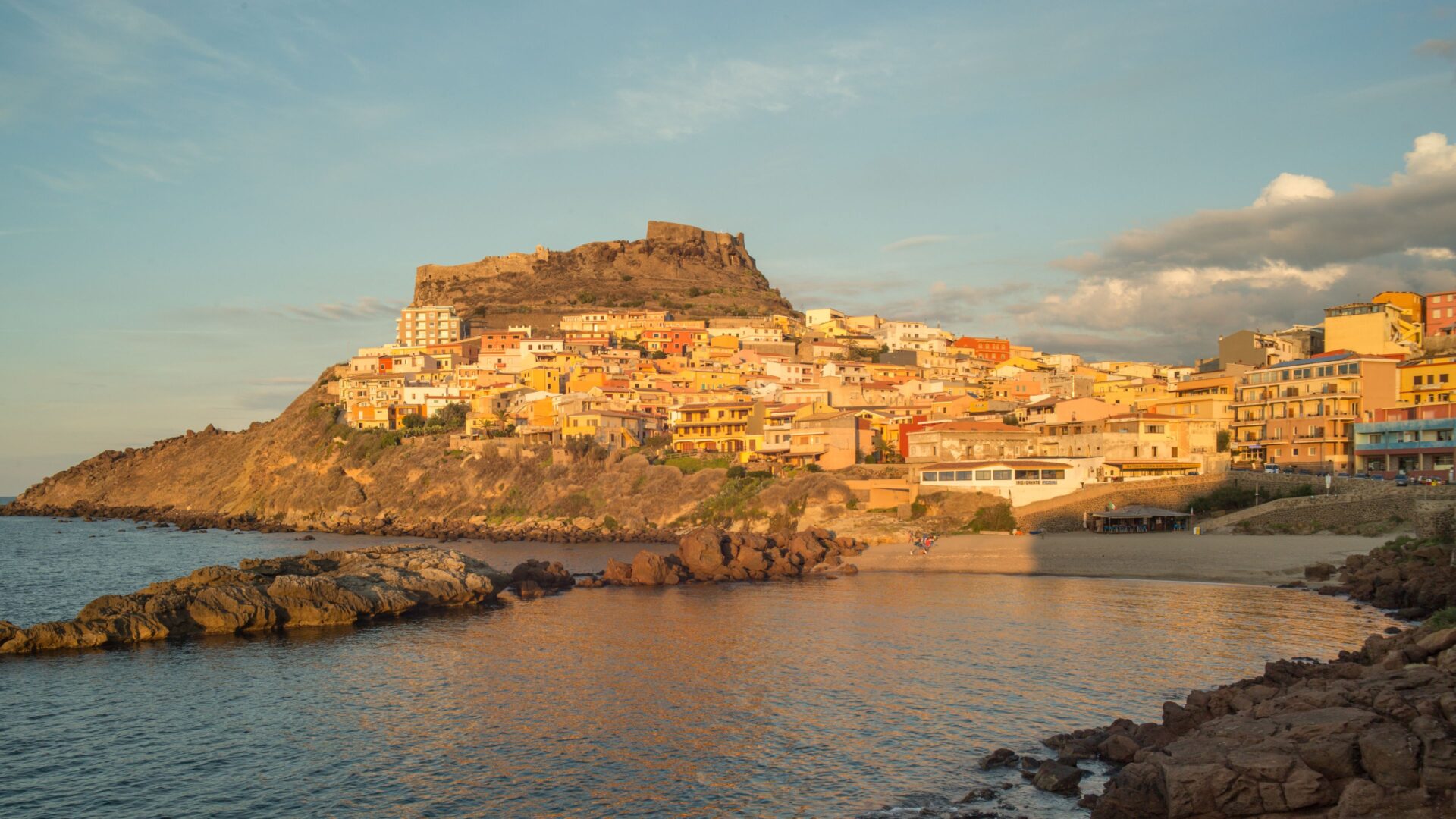 A scenic sunset view of Castelsardo, Sardinia, featuring colorful buildings cascading down a hillside towards the sea.