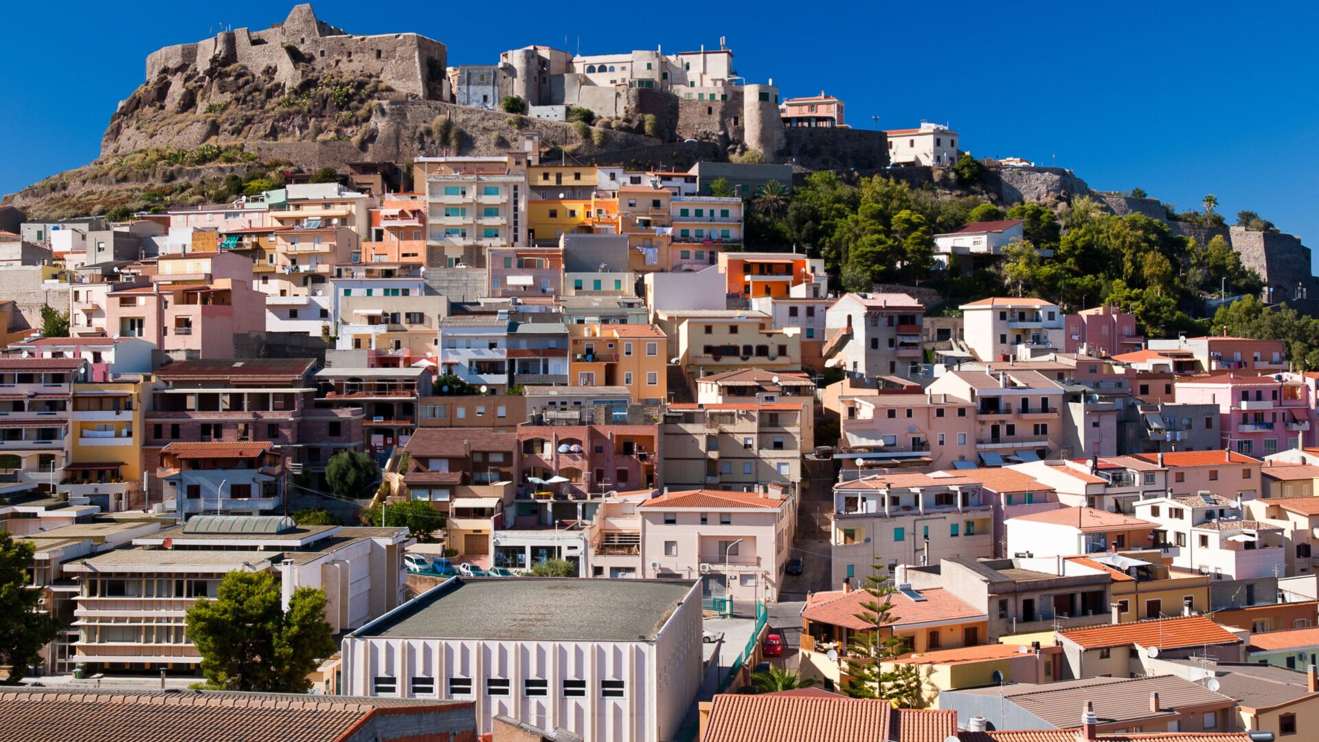  A scenic view of Castelsardo, Sardinia, featuring colorful buildings cascading down a hillside towards the sea, with a medieval castle perched on the hilltop overlooking the town.