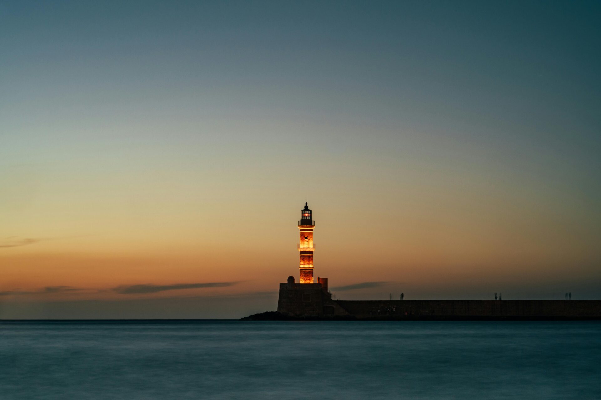 Chania Lighthouse at dusk, with a fiery sky and gentle waves lapping the pier