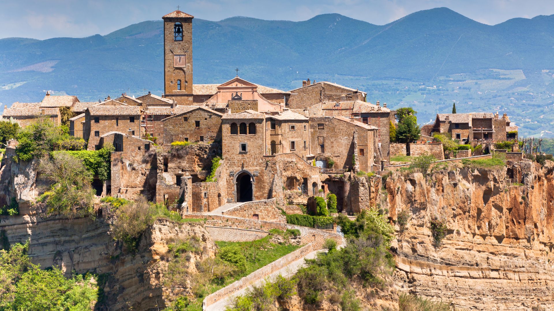 A panoramic view of Civita di Bagnoregio, an ancient Italian town perched atop a cliff, with its medieval buildings and a long bridge leading to it, set against a backdrop of rolling hills.