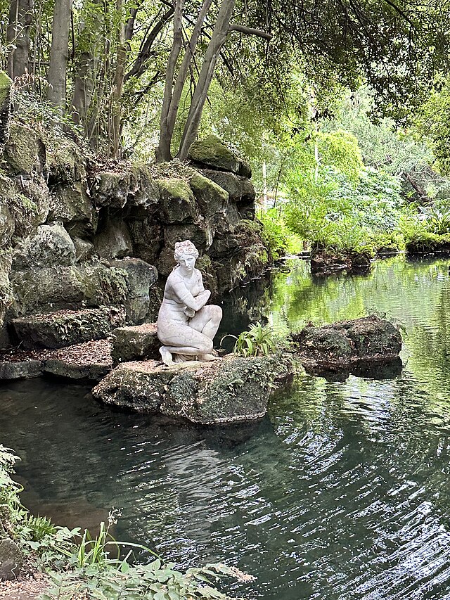 Marble statue of a woman sitting by a peaceful pond surrounded by greenery in the English Garden, Caserta.