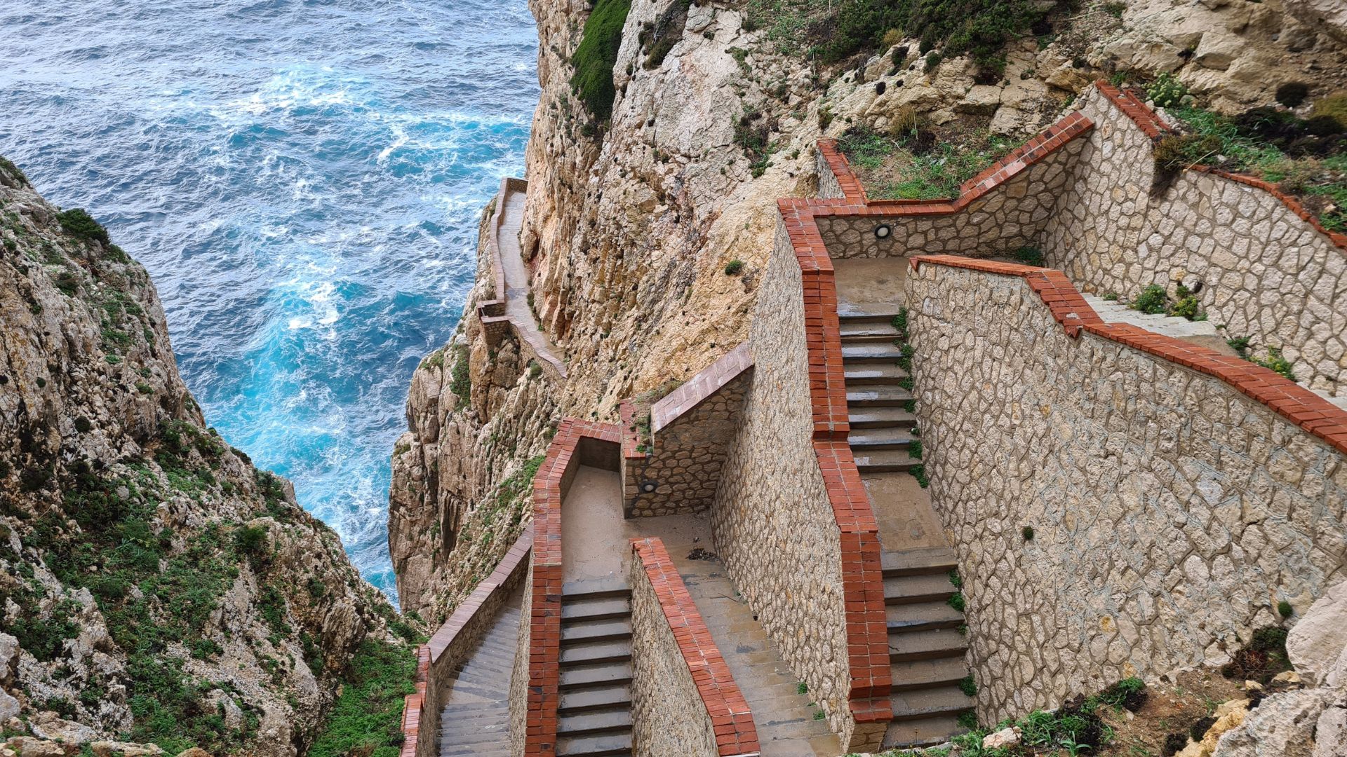 A high-angle view of the Escala del Cabirol, a long, winding staircase with brick-lined stone walls, descending a steep cliff face towards the turquoise waters of the Mediterranean Sea.