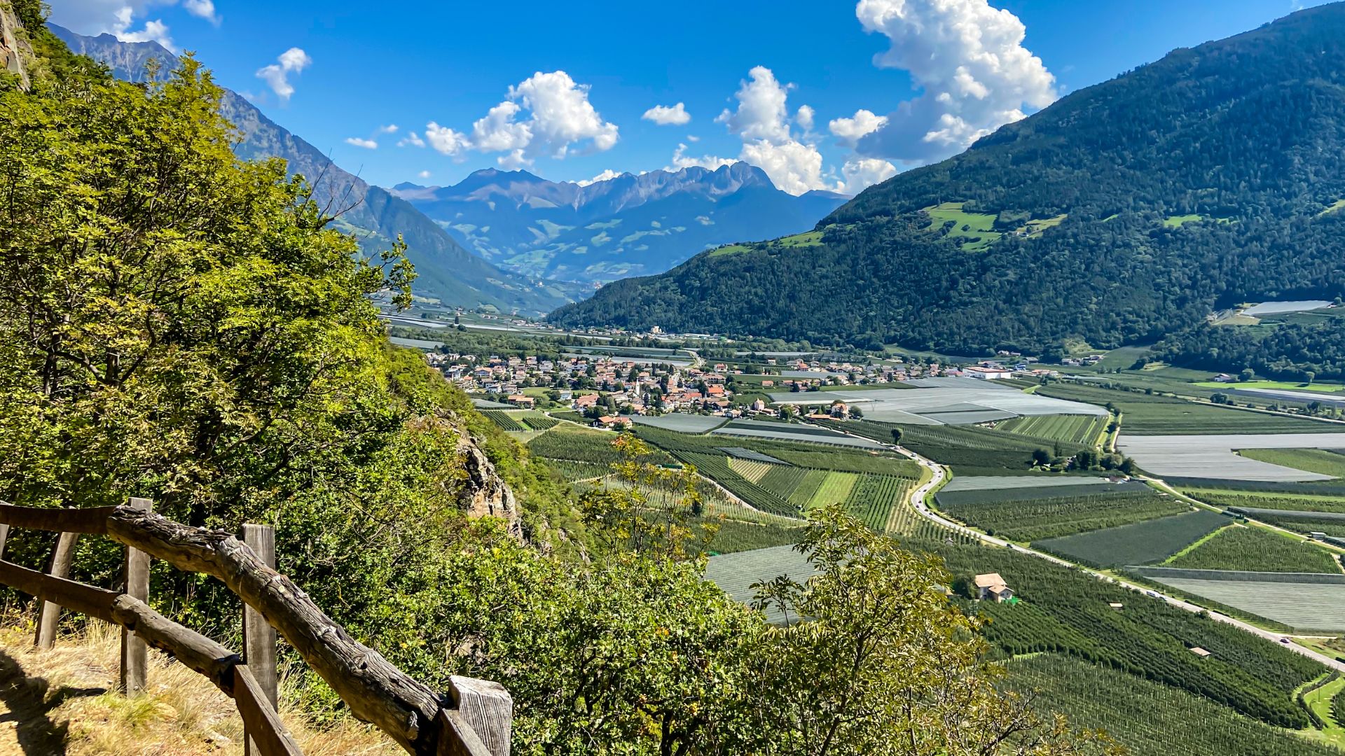 Scenic view of the Etschtal Valley in South Tyrol, Italy, featuring terraced fields, a small village, and surrounding mountains under a partly cloudy sky.