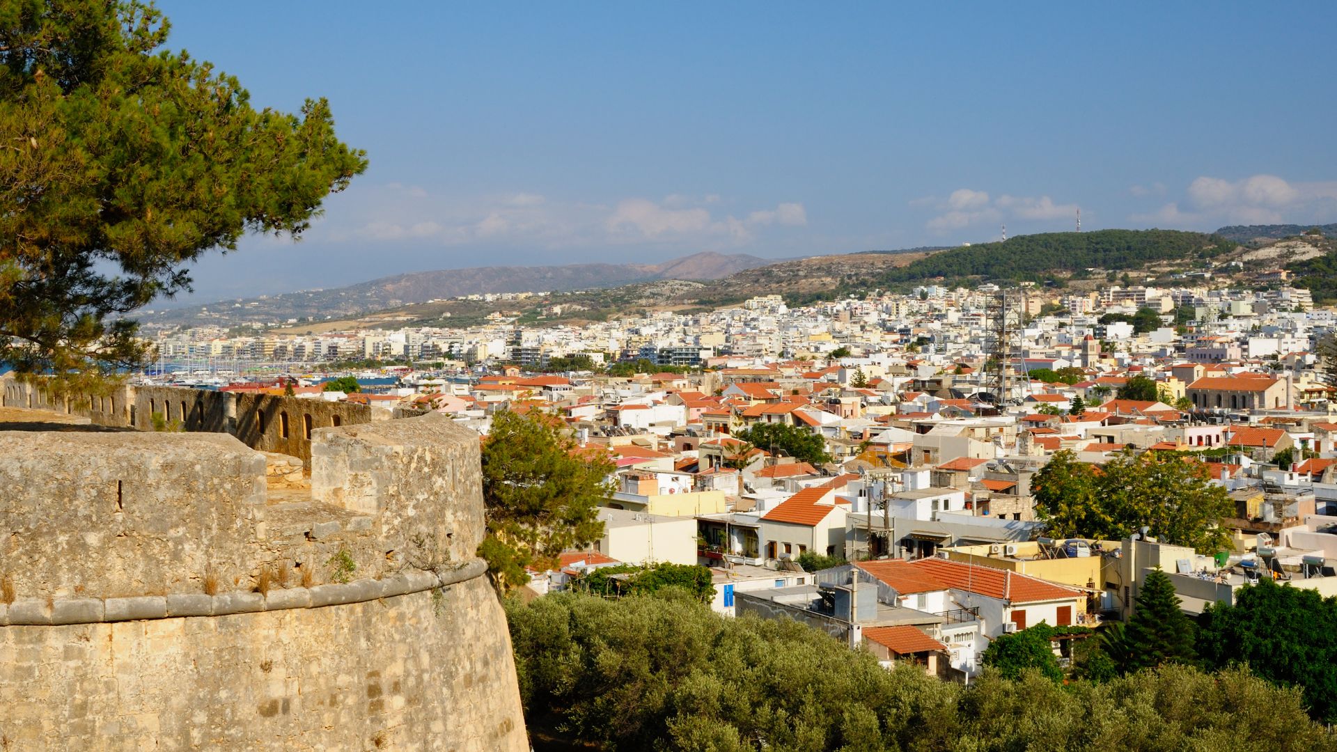 A panoramic view of Rethymno, Crete, from the Fortezza, showing the town's buildings with red-tiled roofs, the coastline, and distant mountains under a clear blue sky.