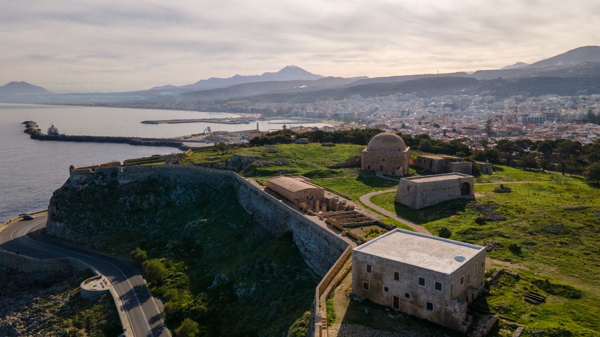 Fortezza of Rethymno, a Venetian-era fortress on the coast of Crete, Greece, featuring stone walls, buildings, and surrounding landscape with the city and mountains in the background.
