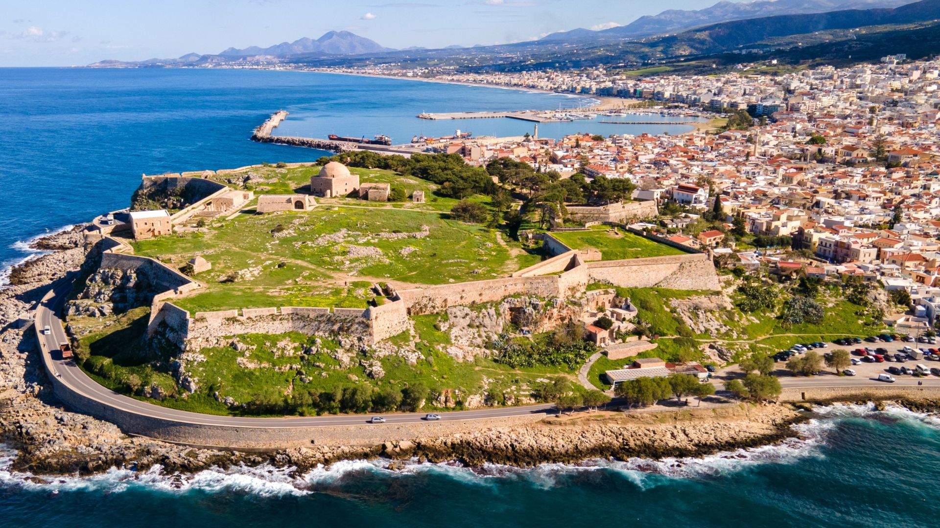 Fortezza of Rethymno, a Venetian-era fortress on the coast of Crete, Greece, featuring stone walls, buildings, and surrounding landscape with the city and mountains in the background.