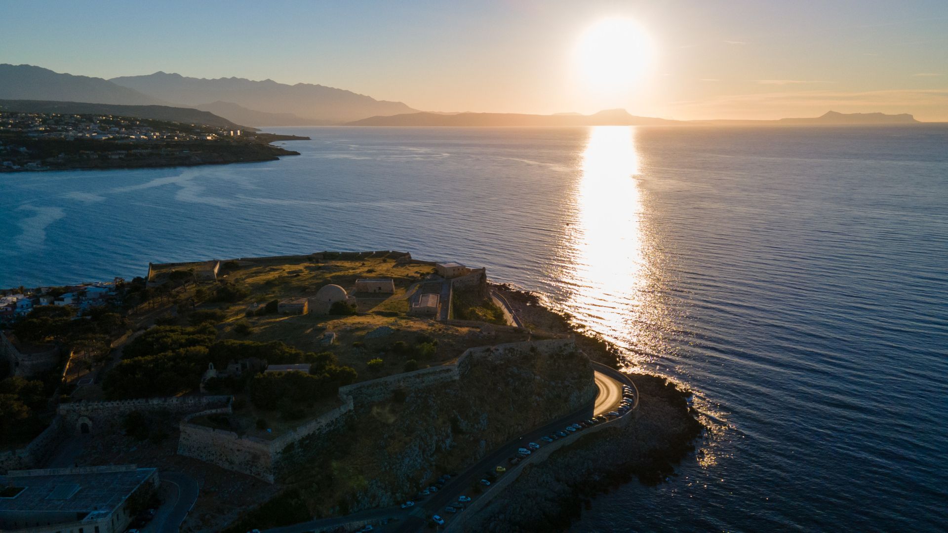 Sunset view of the Fortezza of Rethymno, a Venetian fortress on the island of Crete, Greece, with the sun setting over the sea. The fortress is silhouetted against the orange sky, and the water reflects the light of the setting sun. 