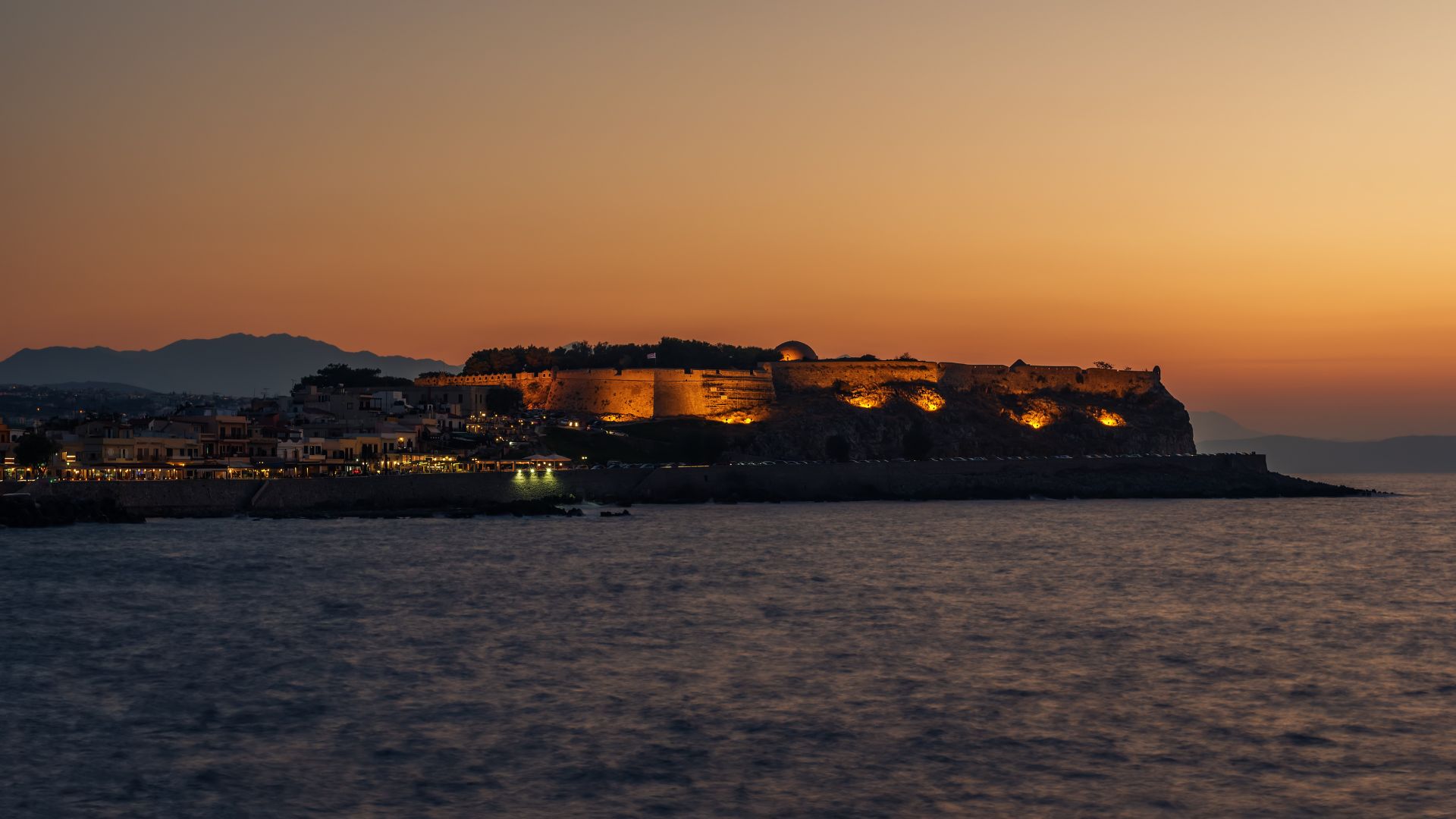 Sunset view of the Fortezza of Rethymno, a Venetian fortress on the island of Crete, Greece, with the sun setting over the sea. The fortress is silhouetted against the orange sky, and the water reflects the light of the setting sun.