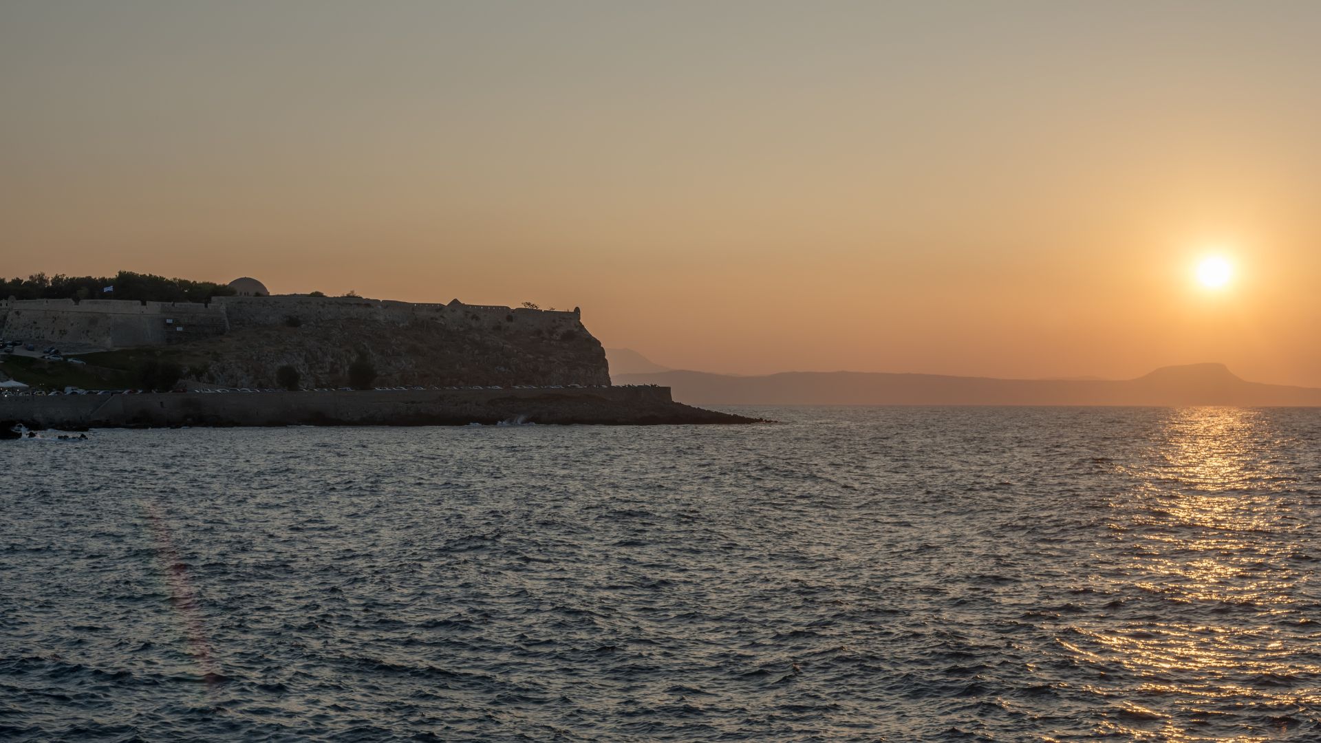 Sunset view of the Fortezza of Rethymno, a Venetian fortress on the island of Crete, Greece, with the sun setting over the sea. The fortress is silhouetted against the orange sky, and the water reflects the light of the setting sun. 