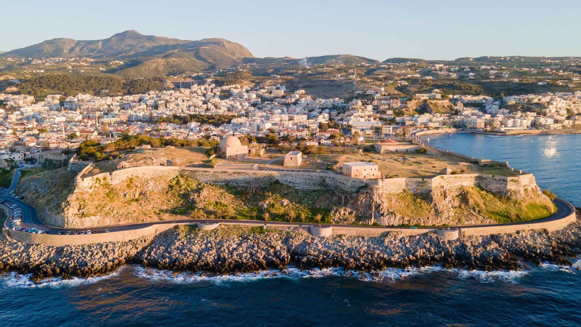 Fortezza of Rethymno, a Venetian-era fortress on the coast of Crete, Greece, featuring stone walls, buildings, and surrounding landscape with the city and mountains in the background.
