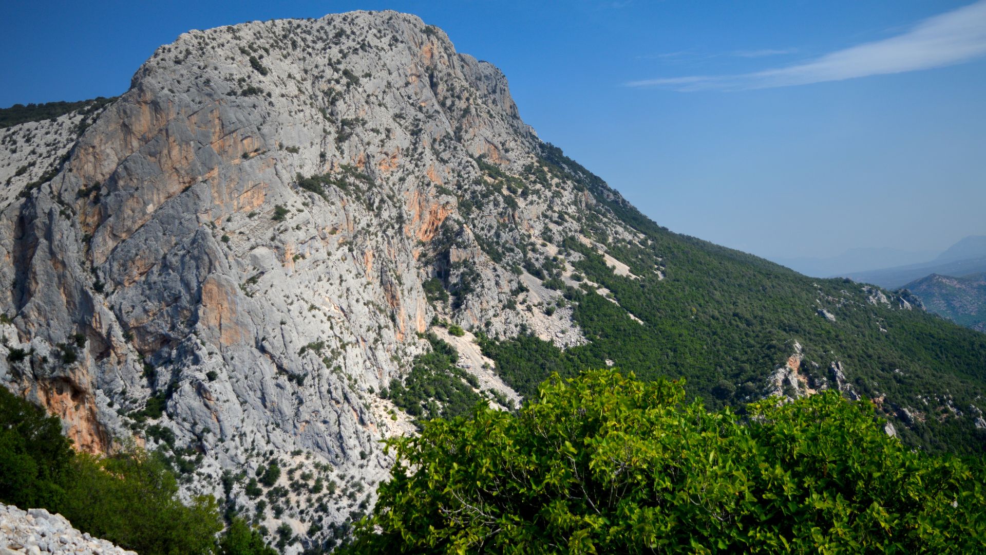 A wide shot of the Gorropu Gorge, a deep canyon with rocky, grey and orange cliffs, partially covered with green vegetation, under a bright sky.