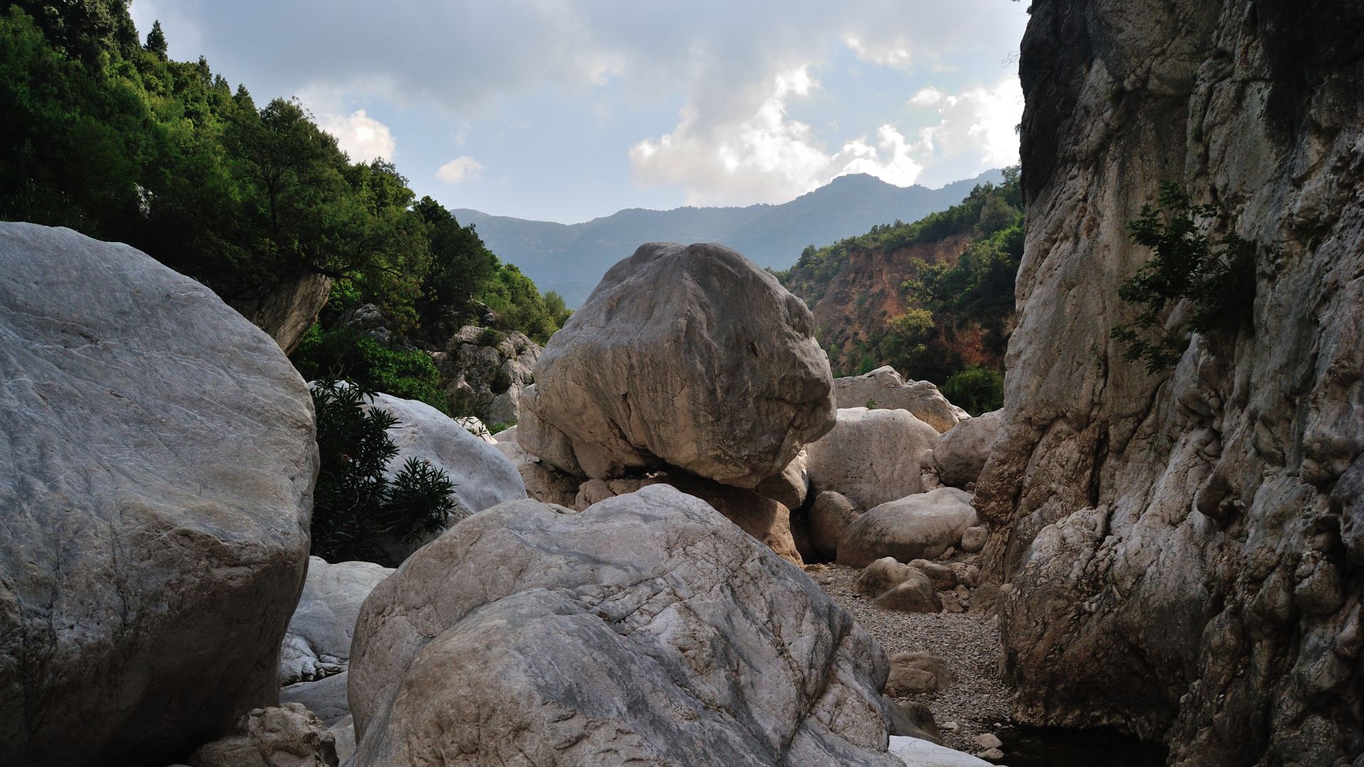 A landscape view of Gola di Su Gorropu, featuring large, smooth rocks in the foreground and middle ground, with steep rock walls on either side and mountains visible in the distance under a cloudy sky.