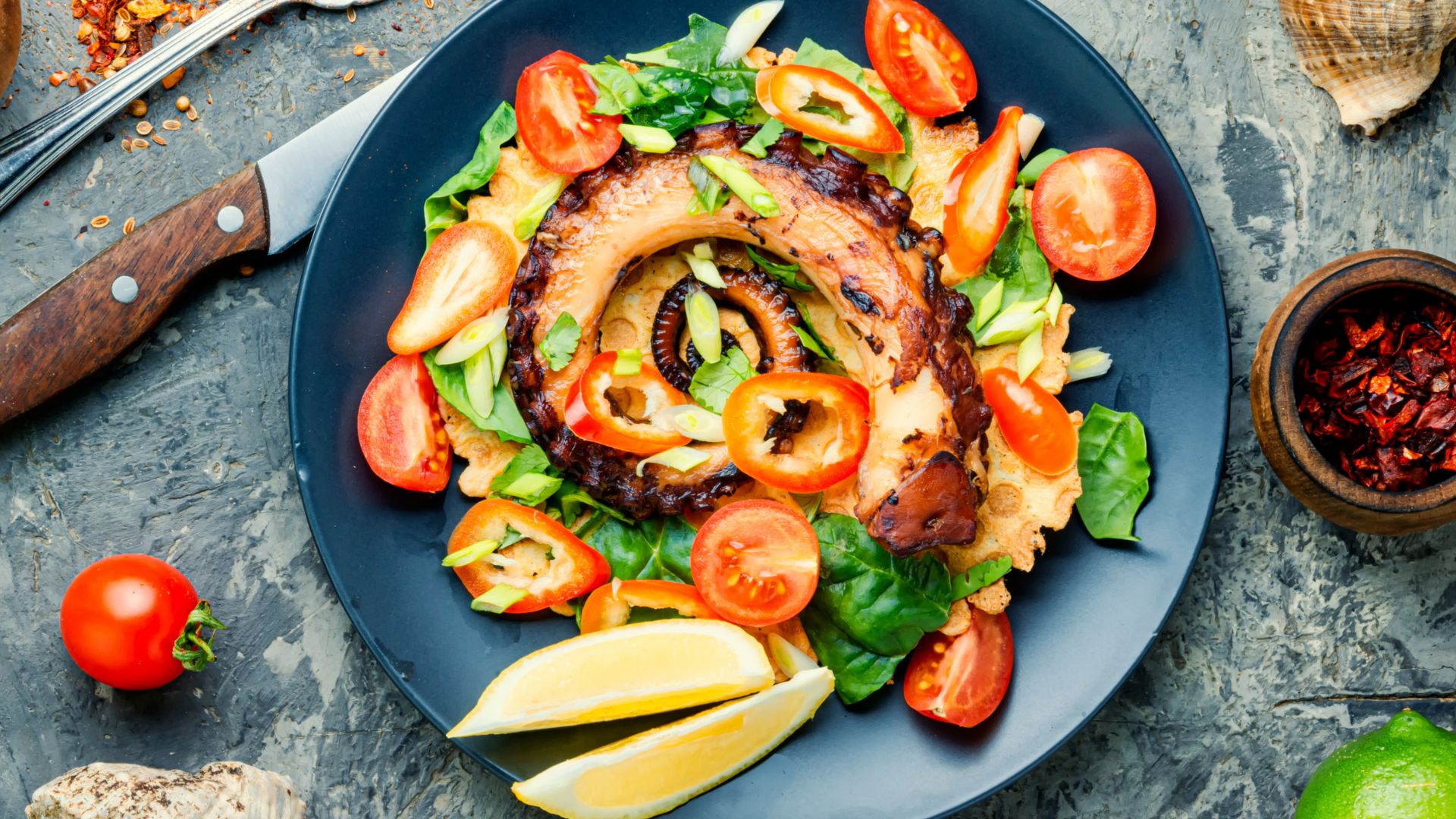 A plate of grilled octopus and tomato salad, garnished with peppers and herbs.