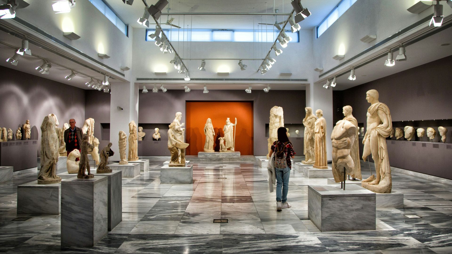 Interior view of the Heraklion Archaeological Museum featuring a visitor among several ancient statues.