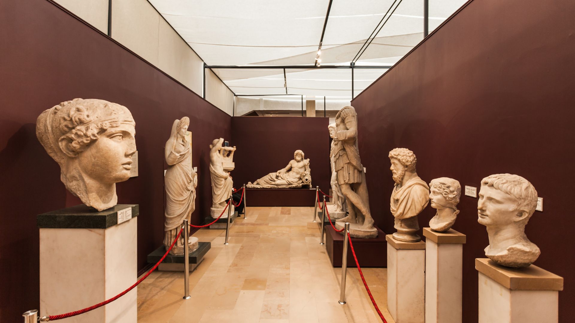 Interior view of the Heraklion Archaeological Museum showcasing a collection of ancient Greek sculptures and busts displayed on pedestals in a well-lit gallery.